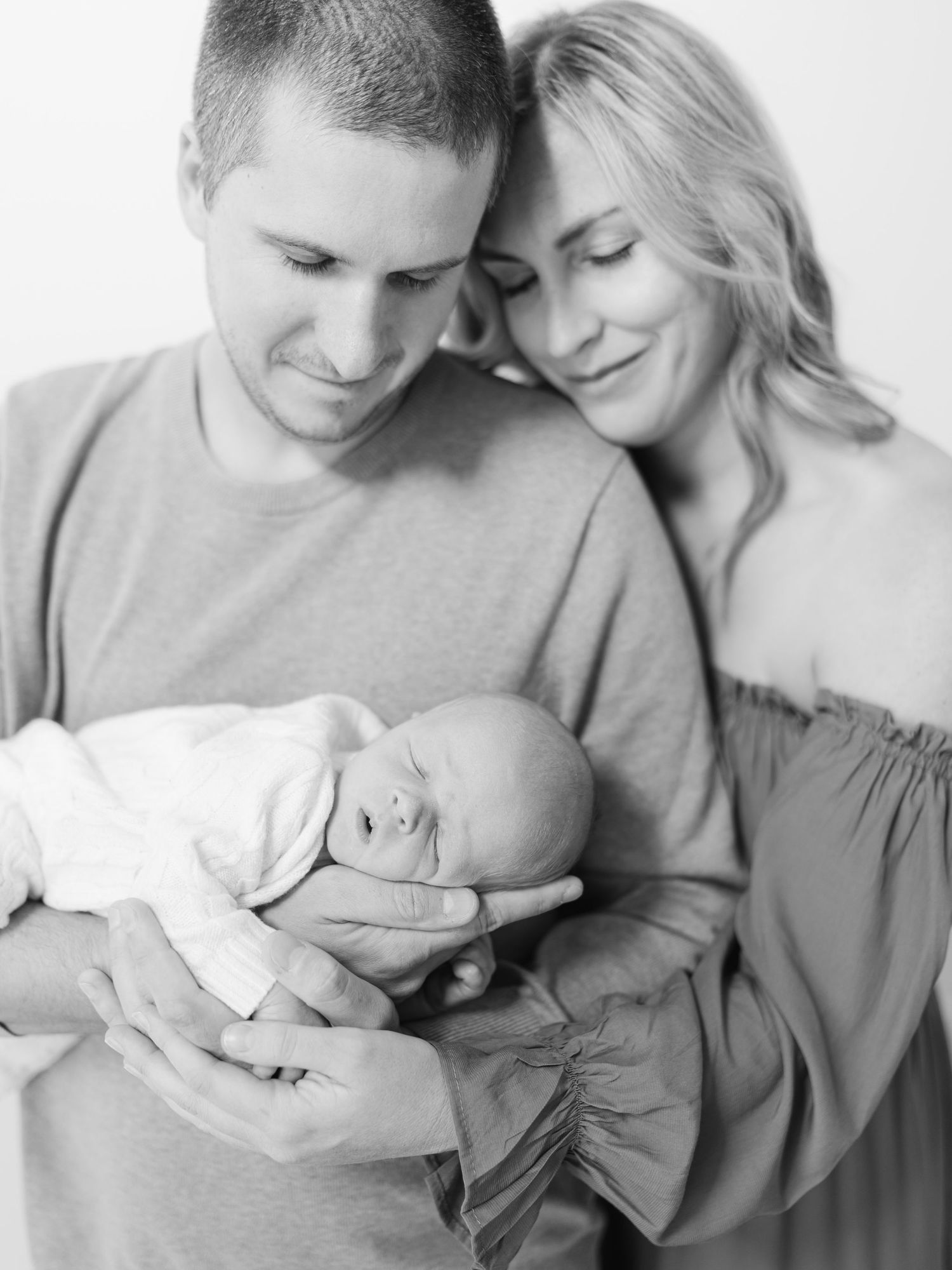 Black and white newborn photo of parents tenderly cradling their sleeping baby, capturing a serene family portrait moment.