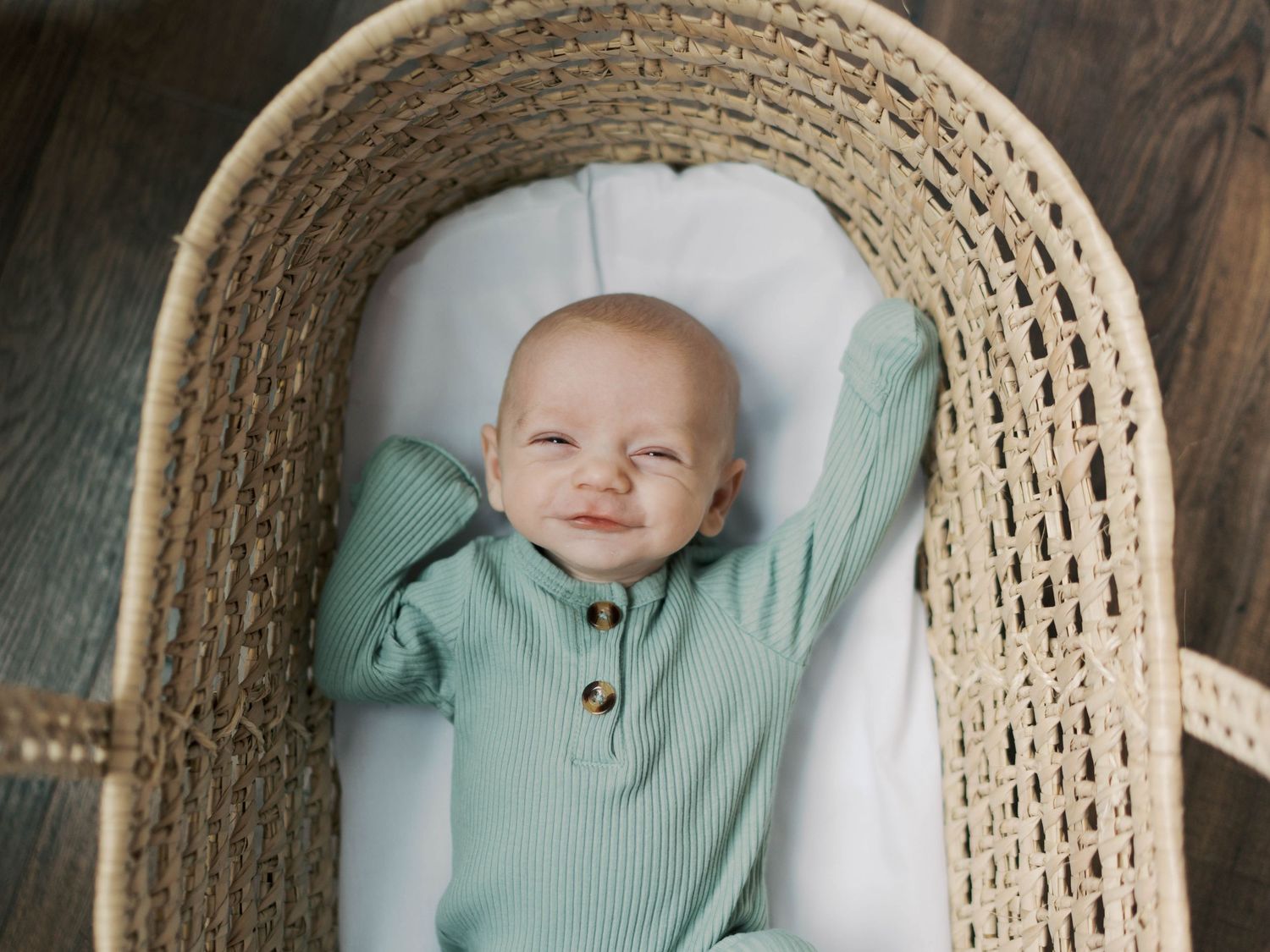 Smiling baby in green ribbed onesie lying in a woven Moses basket on a dark wood floor.