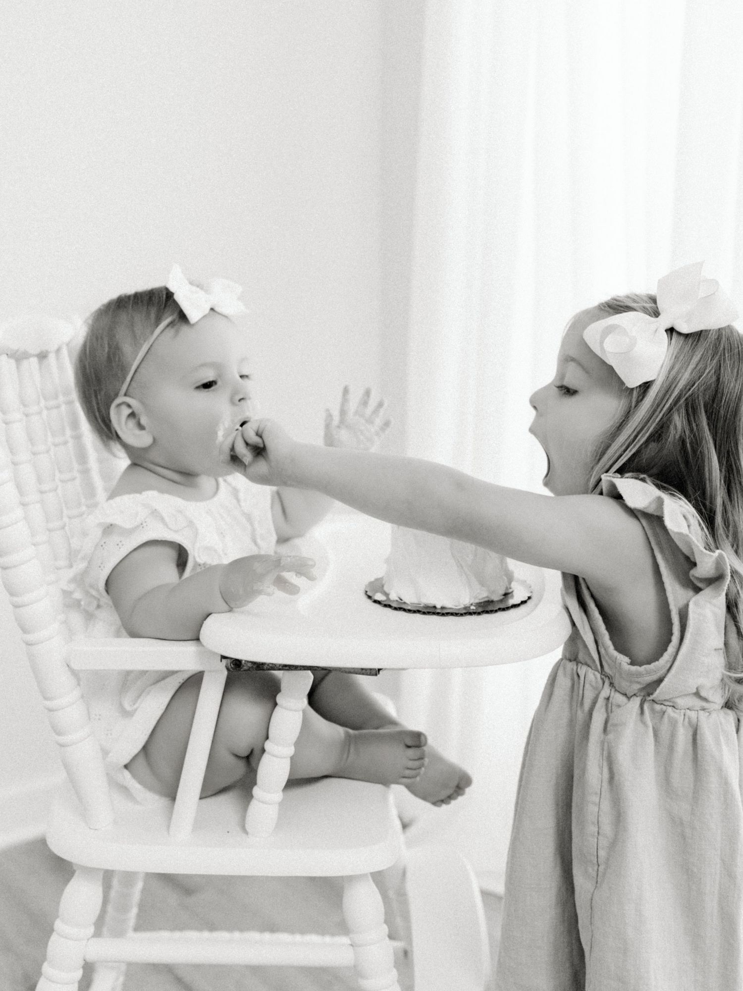 Photo of a toddler in high chair being fed cake by her older sister, both wearing hair bows after visiting Toy Stores in Raleigh