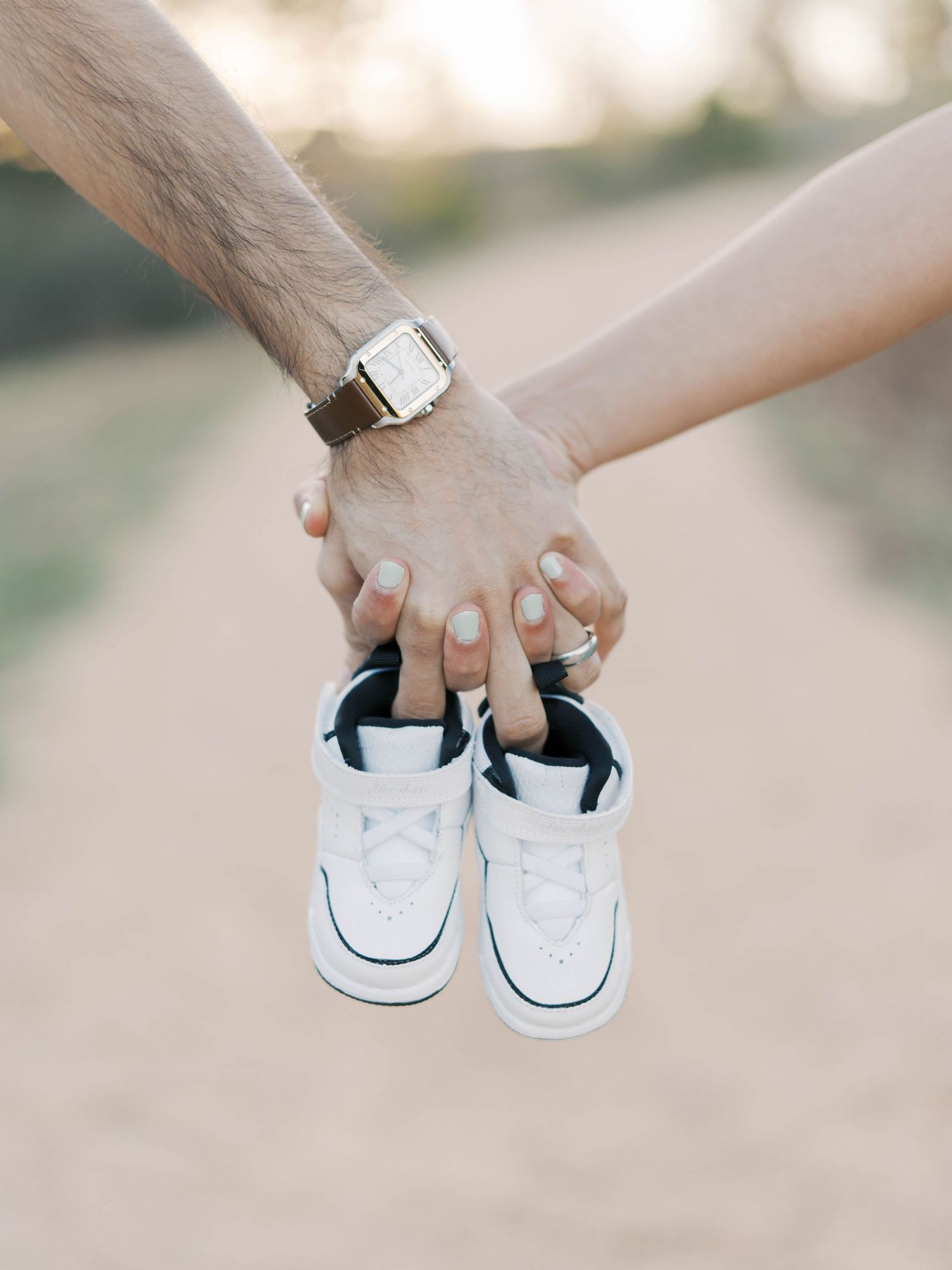 Close up of couple's hands holding tiny white baby shoes against dirt path background.