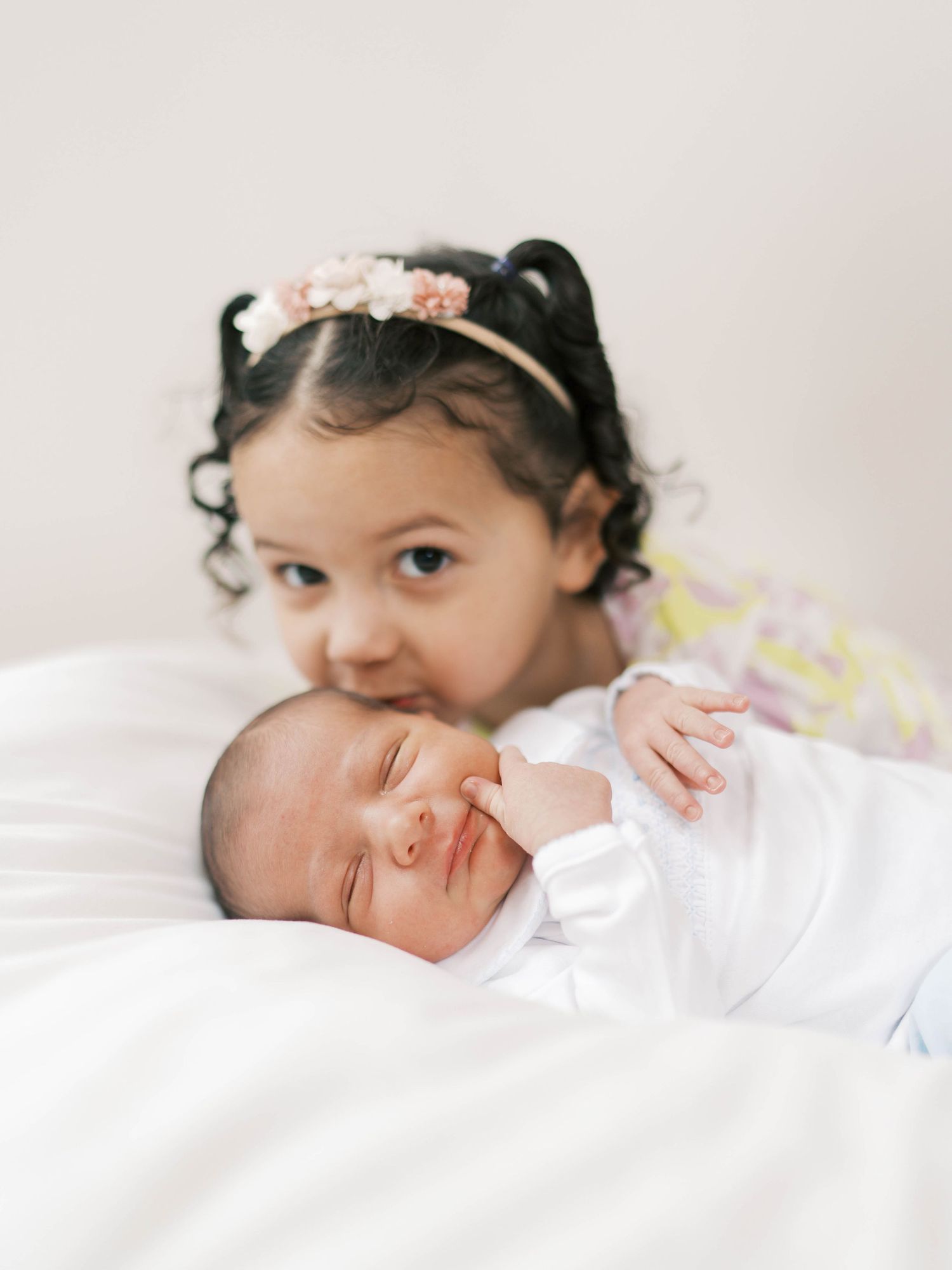 Toddler girl with floral headband lovingly poses with sleeping newborn baby sibling on white bed.