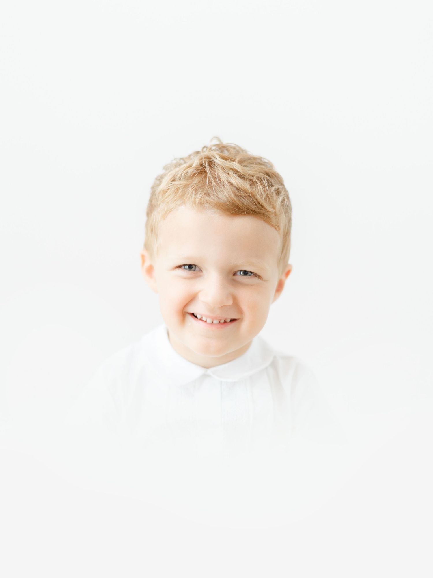 Smiling young blonde boy in a white shirt poses against a bright white background for a portrait photo.