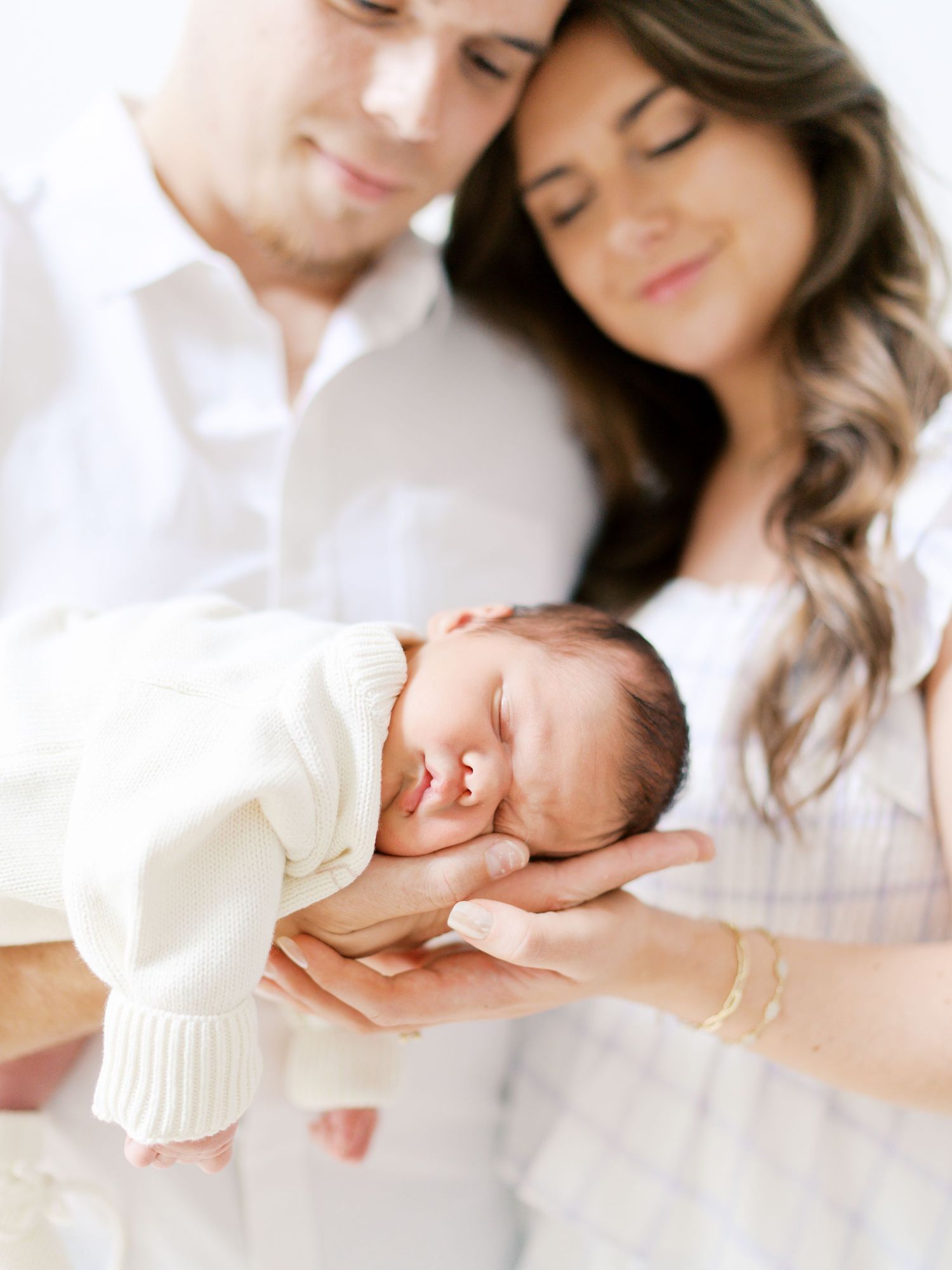 Parents tenderly hold sleeping newborn baby dressed in white cream outfit close to their faces.