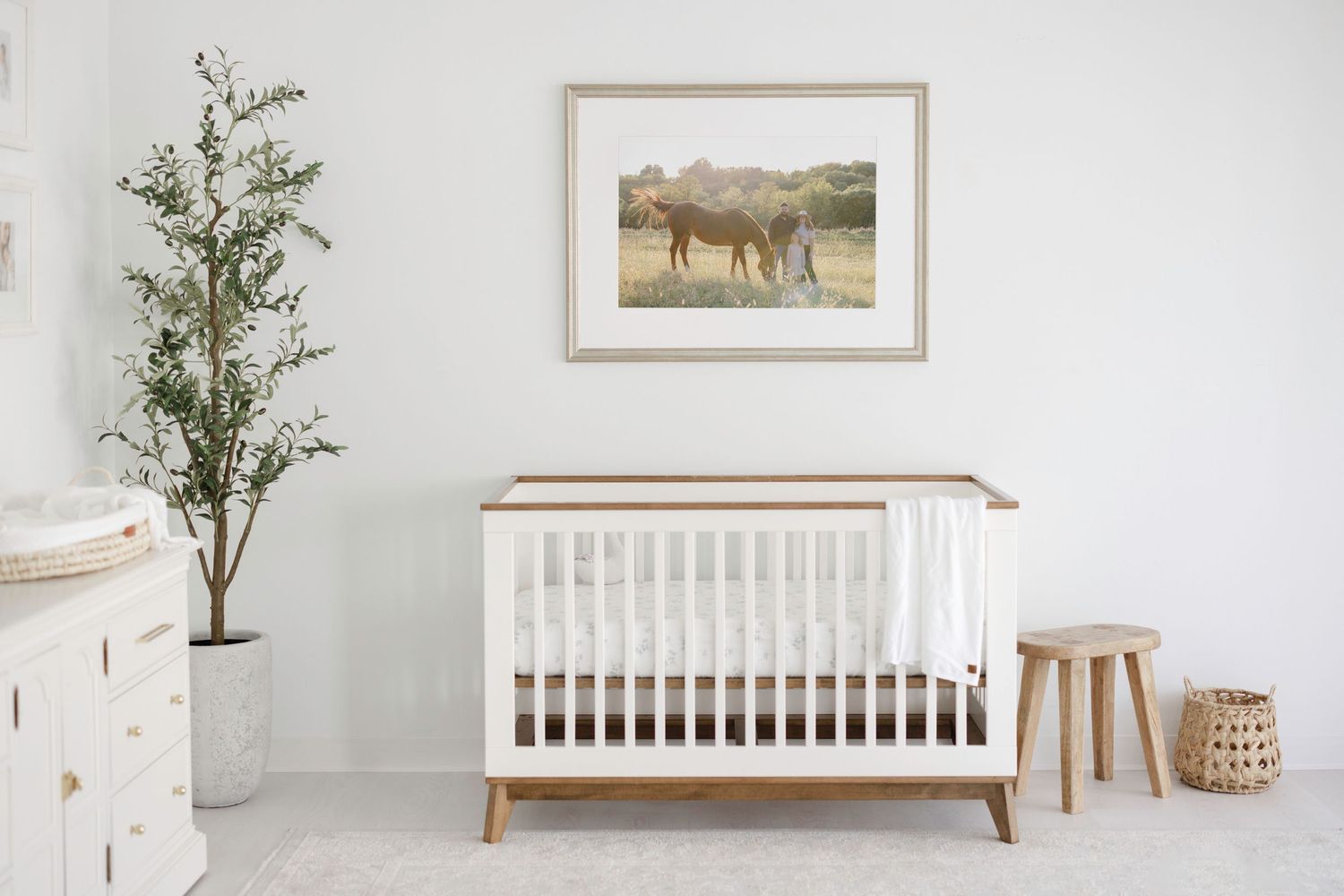 White nursery with a wooden crib, framed horse print, potted olive tree, and natural wood accents on a light gray rug.