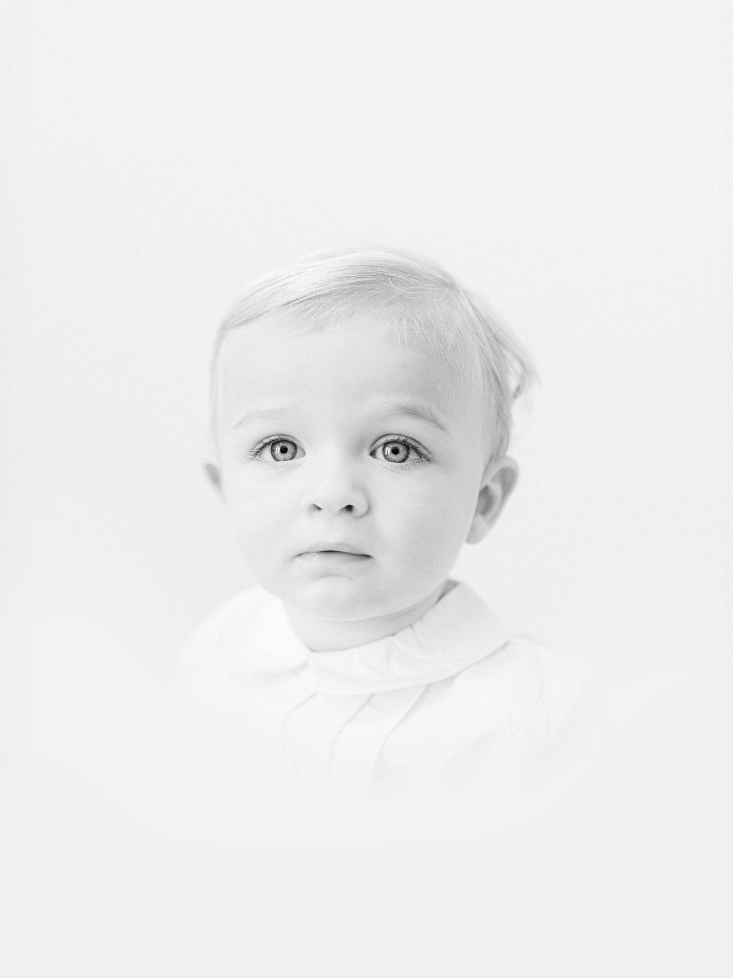 Black and white portrait of an adorable baby with light hair and expressive eyes wearing a white collar shirt.