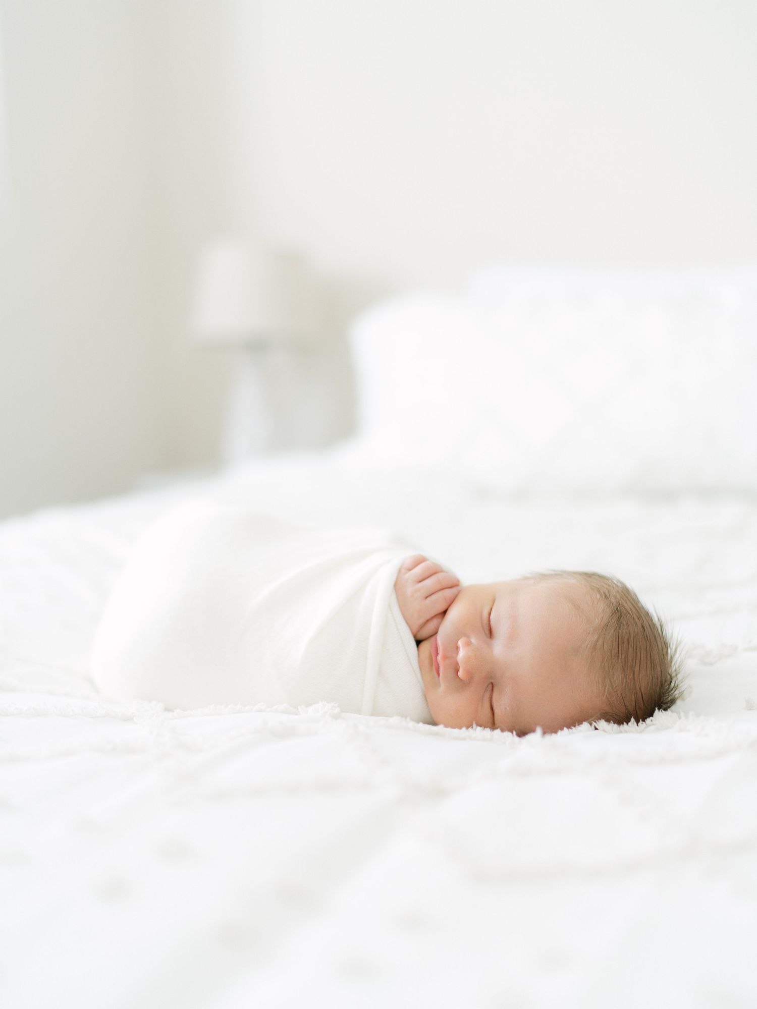 Peaceful newborn baby swaddled in white blanket sleeping on a soft white bed in a bright, airy room.