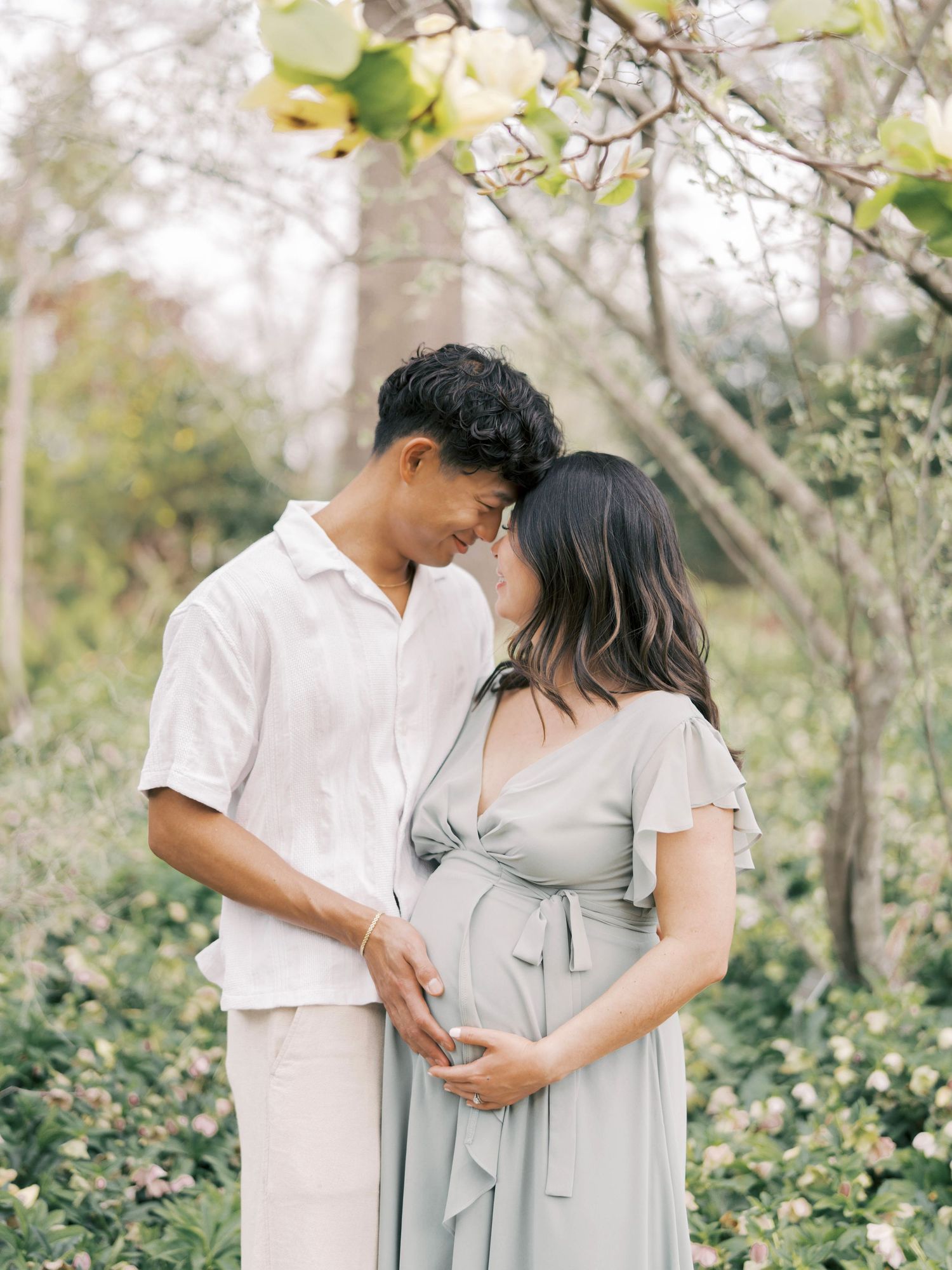 A tender maternity moment captured beneath blooming spring trees in soft natural light.