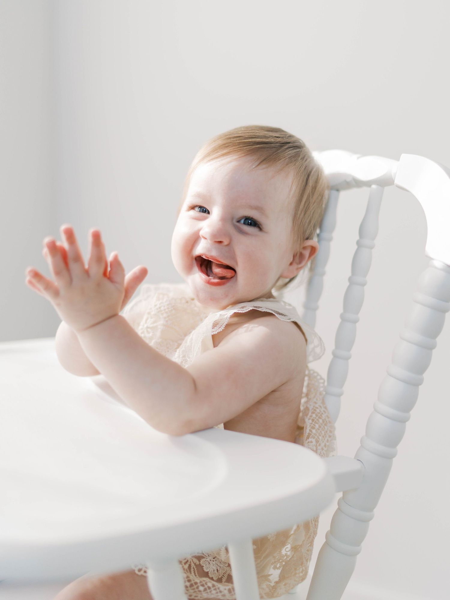 Joyful baby in cream dress clapping while sitting in white high chair.