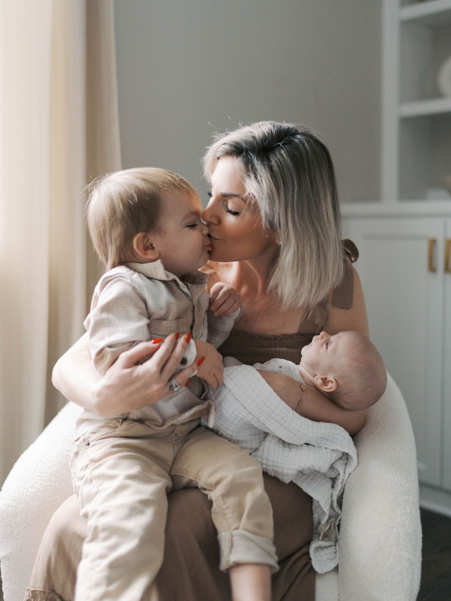 A mother kisses her toddler while cradling a newborn baby in a cozy white chair.
