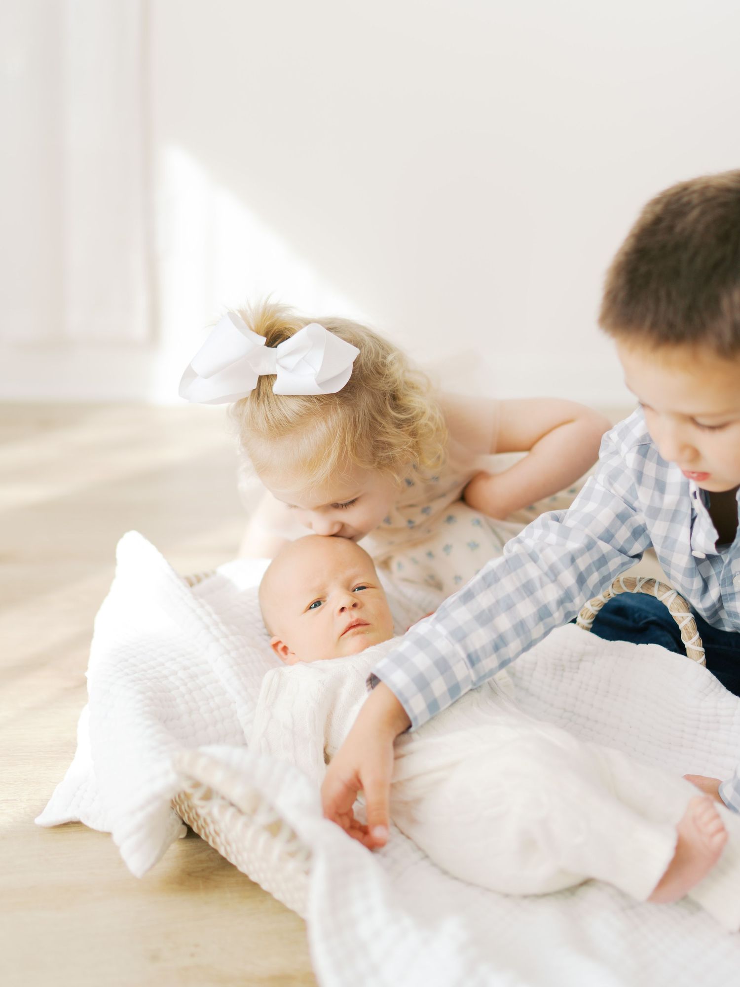 Two toddlers lovingly look at a newborn baby wrapped in white muslin in a wicker basket on a light wood floor.