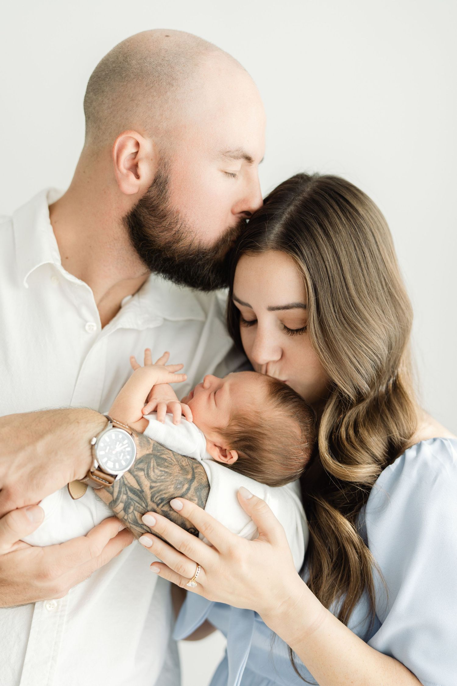 Bald bearded father and brunette mother tenderly kissing their newborn baby against a white background.