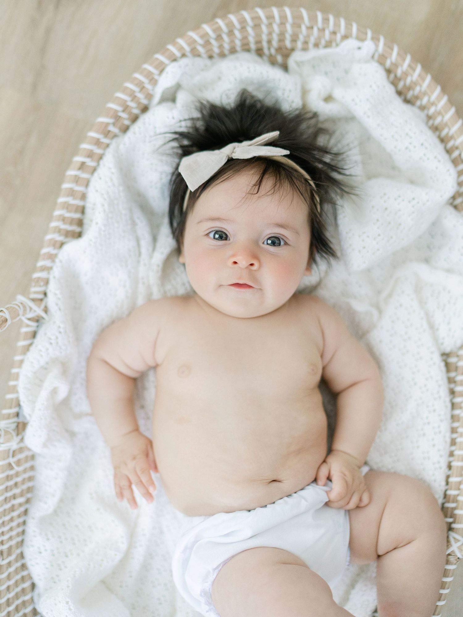 Adorable baby girl with dark hair and bow headband lying in a wicker Moses basket on a white blanket.