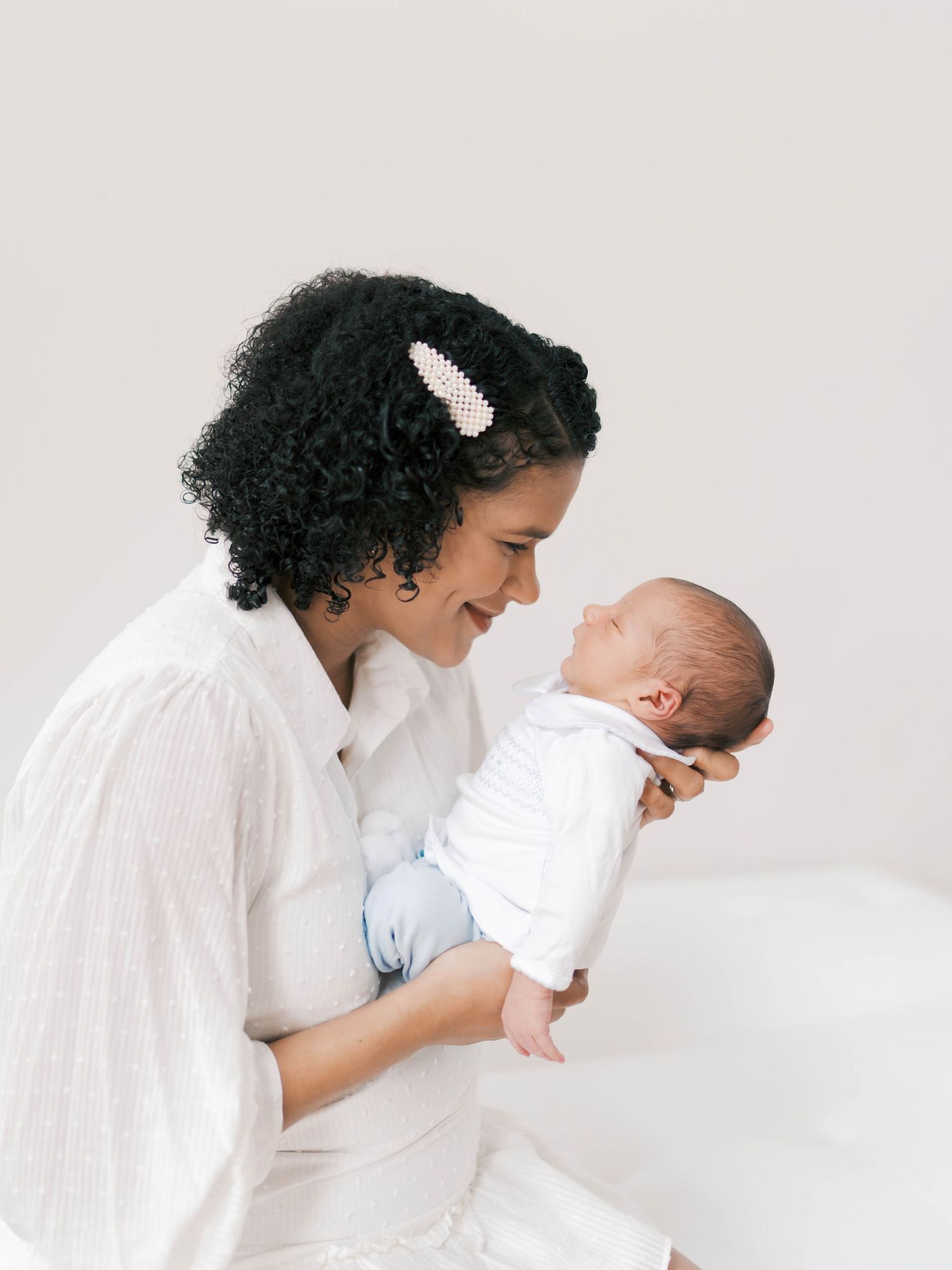 A mother with curly hair smiles tenderly at her newborn baby, both dressed in white against a soft background.