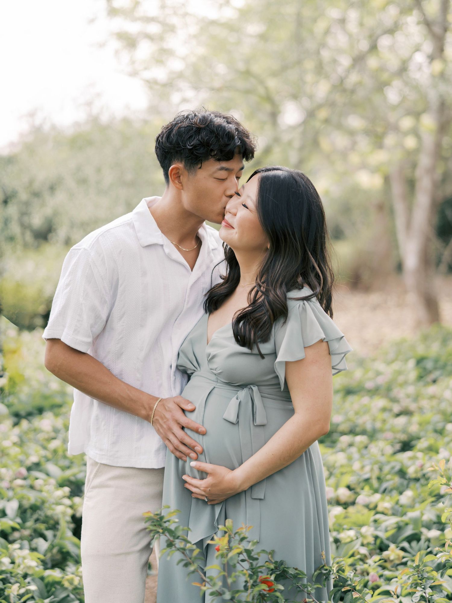 A couple in white and sage green outfits share intimate moments in a lush garden setting during a maternity photoshoot.
