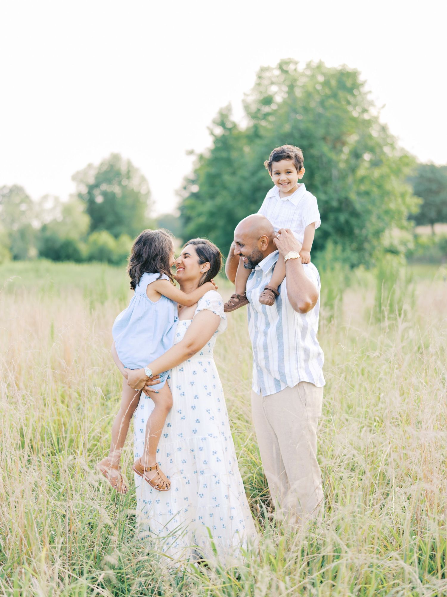 A family in white outfits embraces and plays together in a sunlit summer meadow with tall grass.