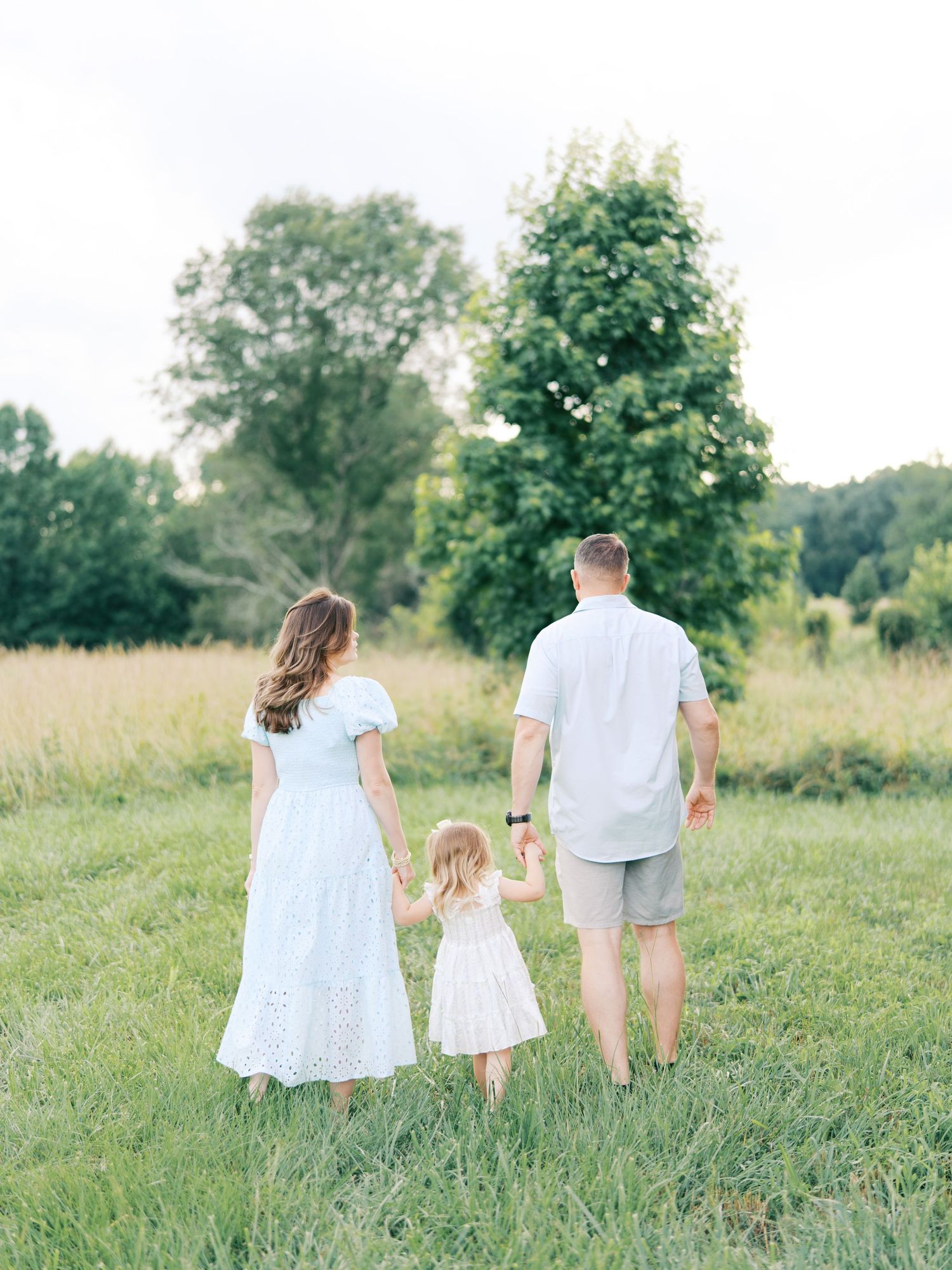 A family walking together through a sunlit meadow wearing white and beige outfits, viewed from behind.