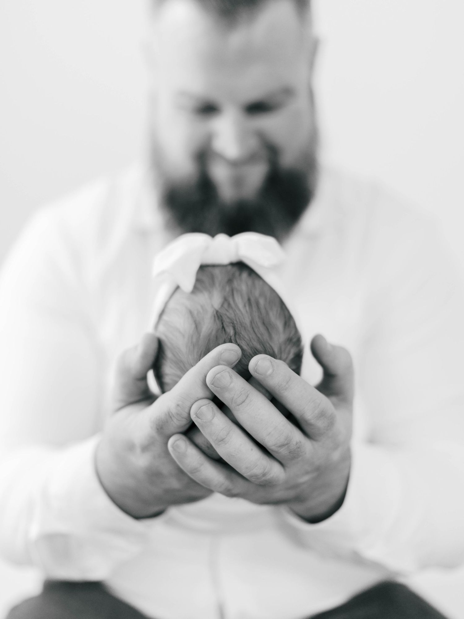 Black and white photo of a bearded father tenderly cradling his newborn baby in both hands.