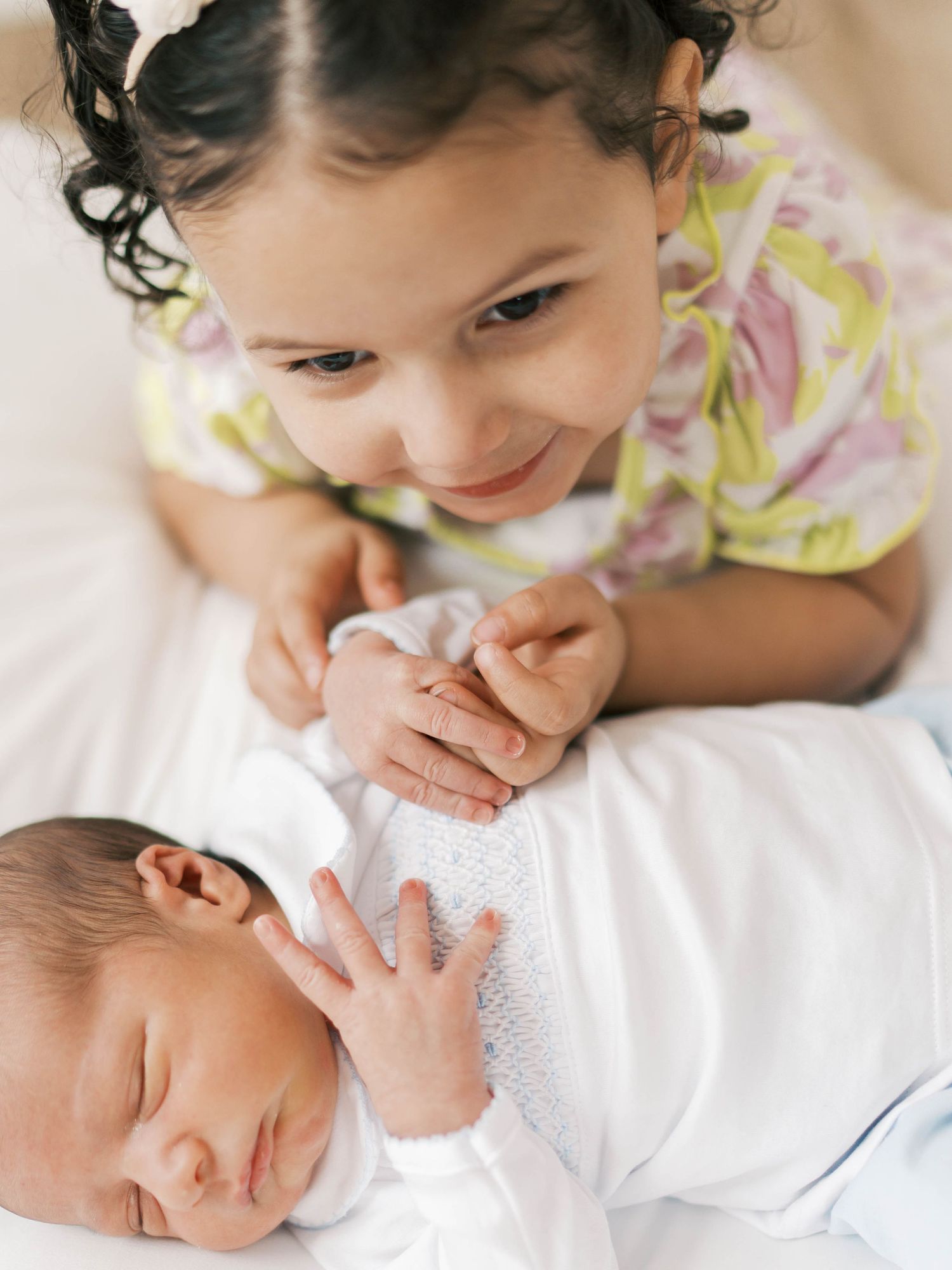 Young girl smiling tenderly at sleeping newborn baby sibling, holding hands in a sweet bonding moment.