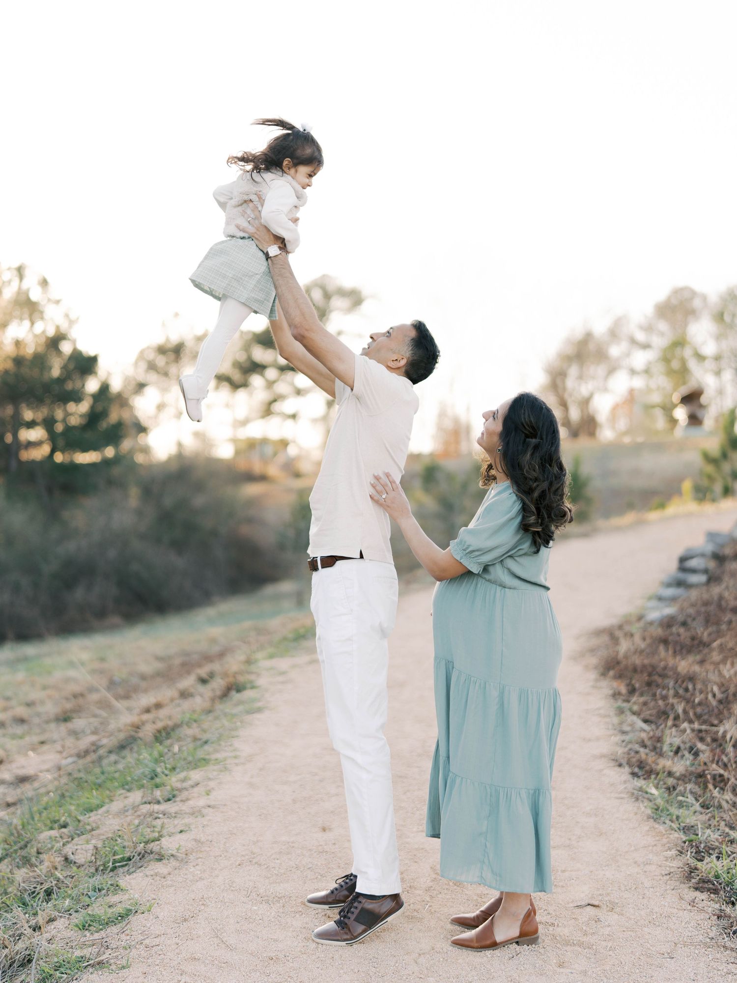 Parent lifts small child in the air while pregnant mother watches on scenic outdoor trail.