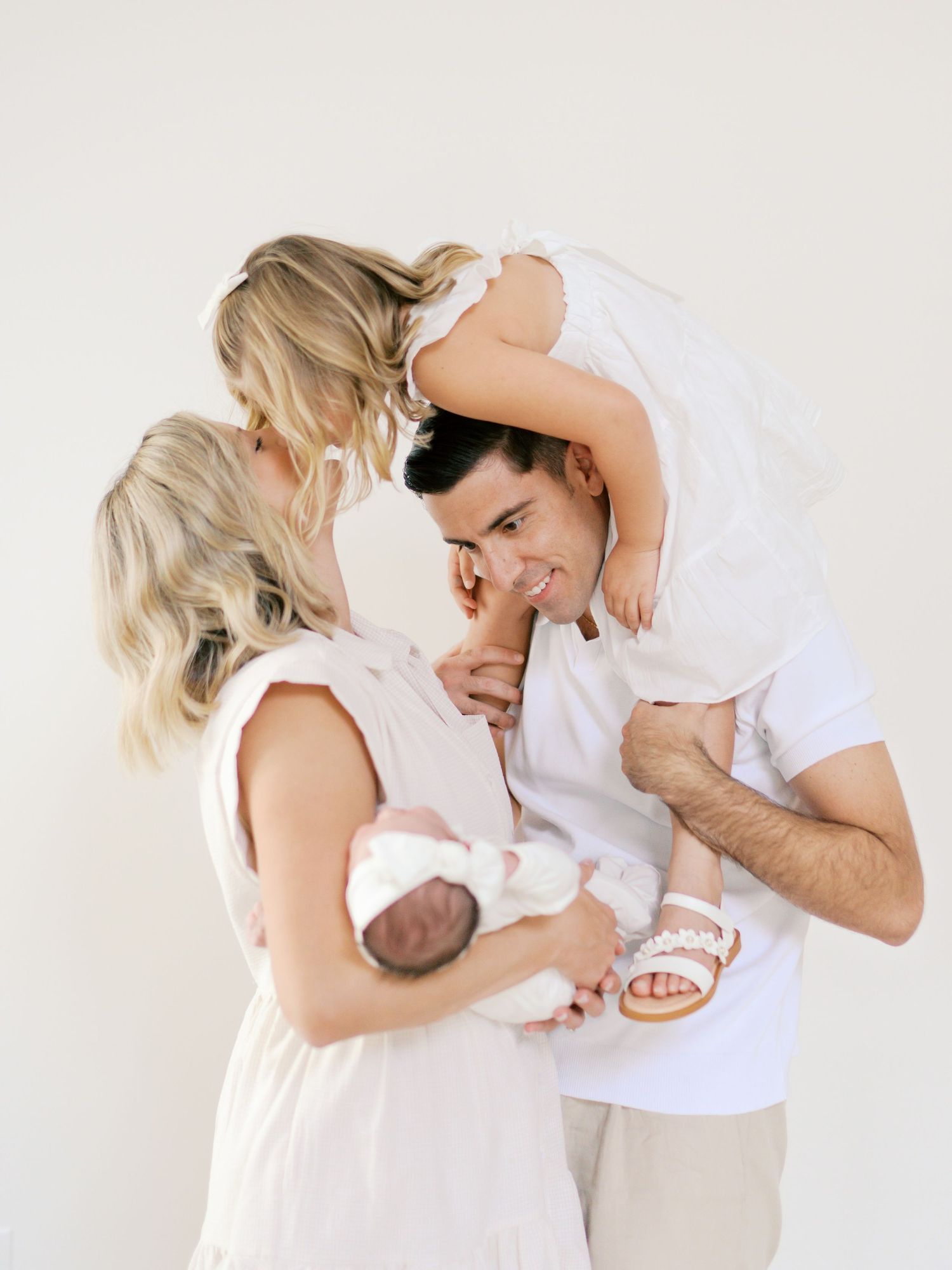 A young family in white outfits shares a tender moment, with mom holding a newborn and dad carrying a toddler.