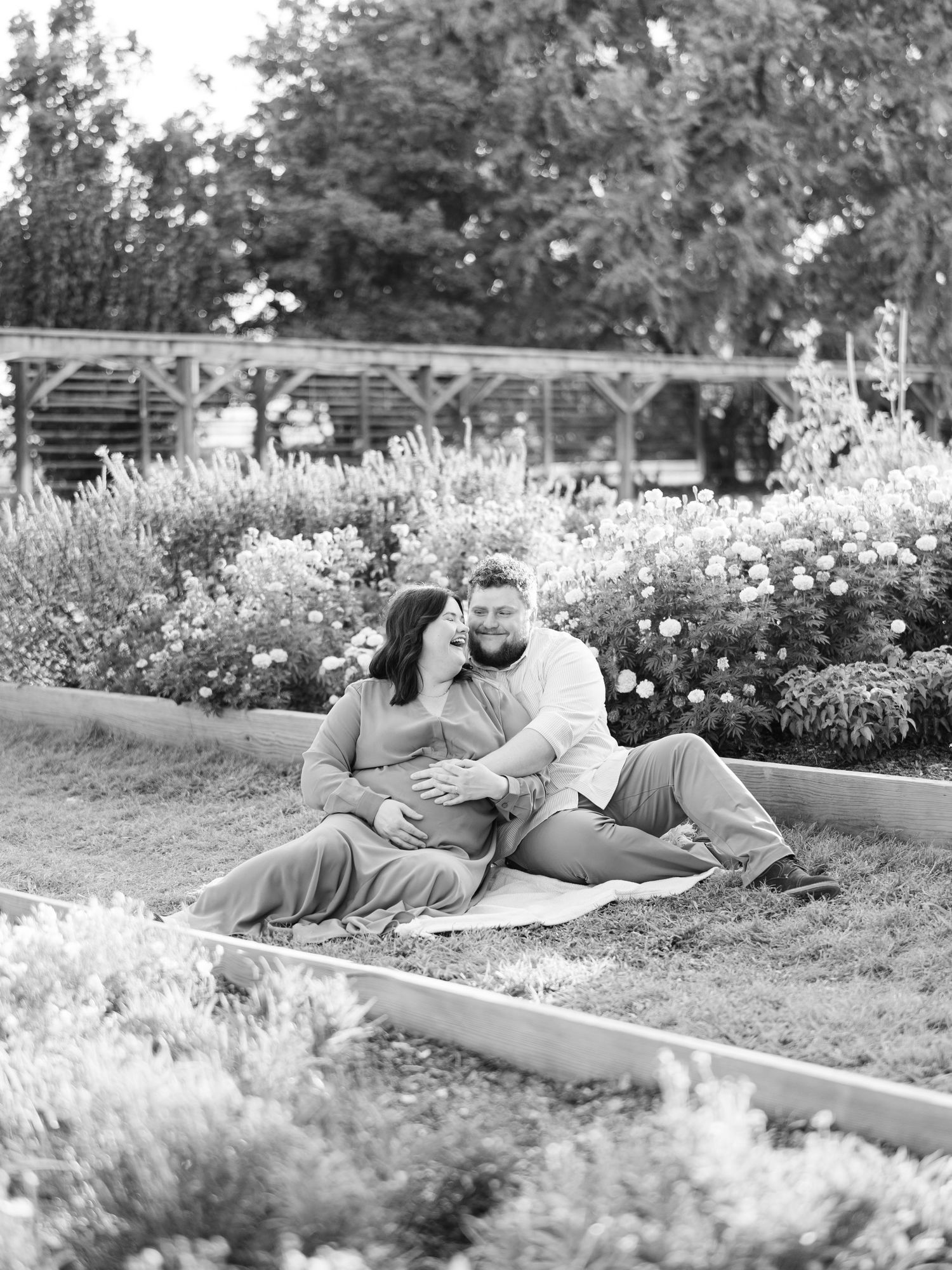Black and white maternity photo of a couple sitting in a lush garden, the man kissing his pregnant partner's head.