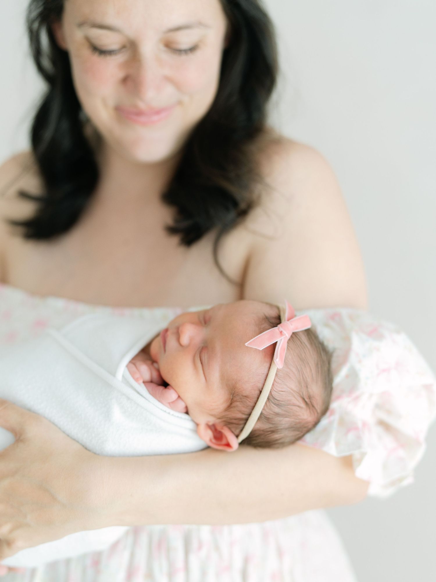 A mother in a floral dress tenderly holds her sleeping newborn baby girl wearing a pink bow headband.