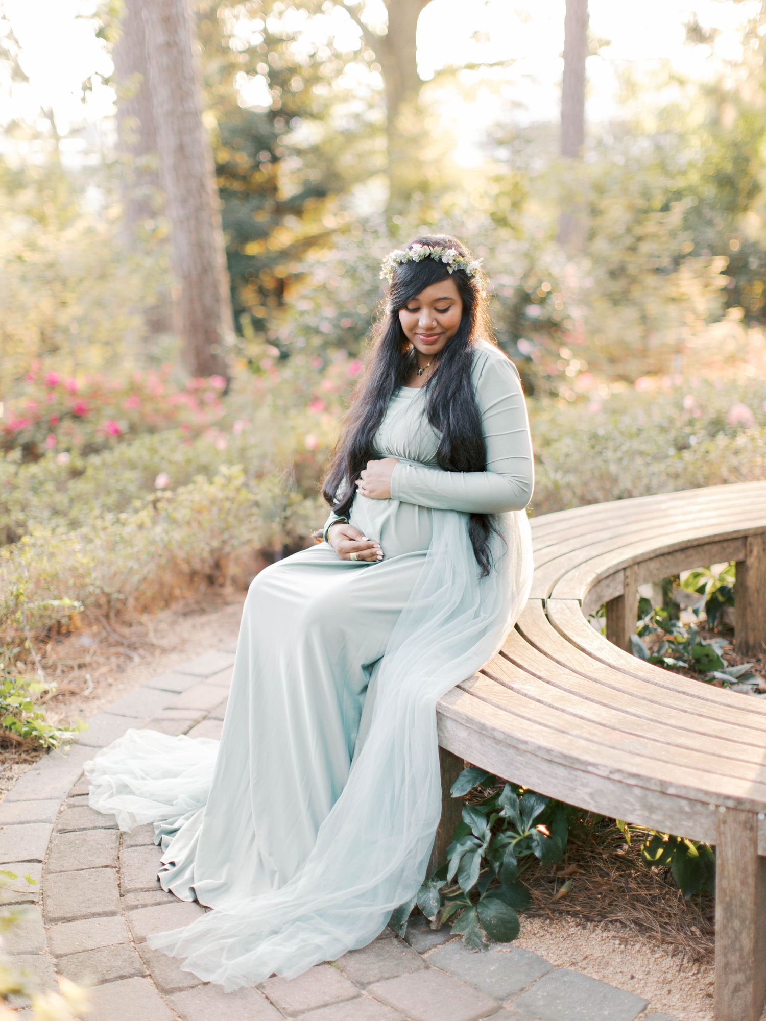 Expectant mother in pale blue gown sits on curved garden bench surrounded by greenery.