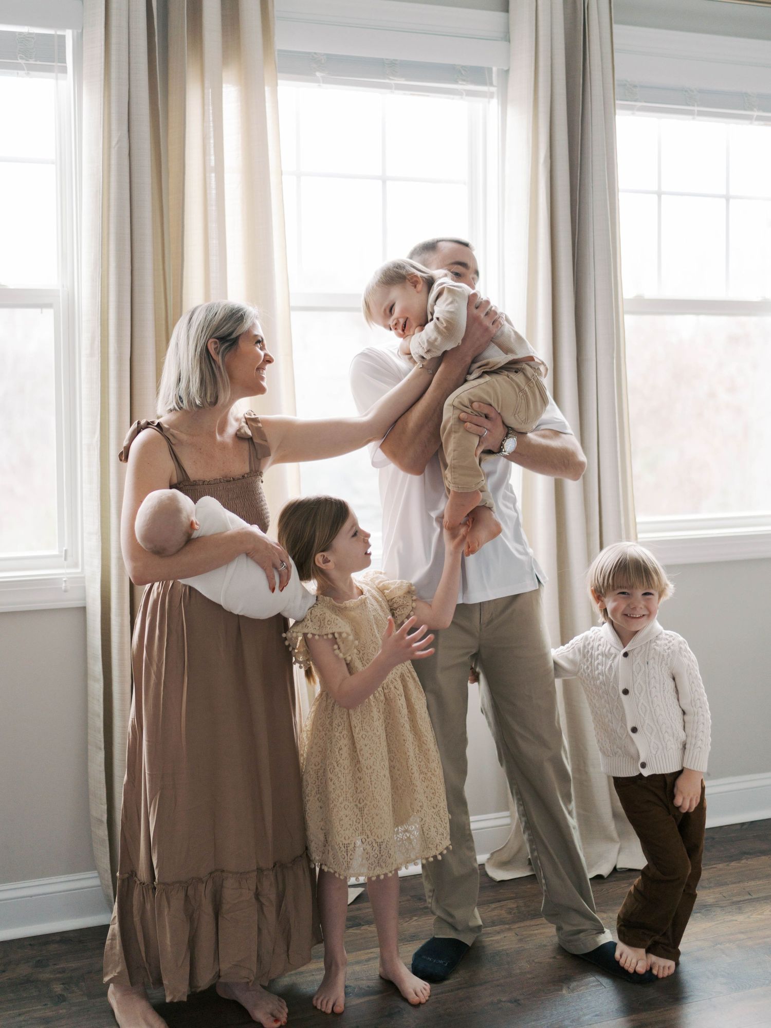 A joyful family of six poses by bright windows, parents lifting a baby while children gather around in neutral tones.