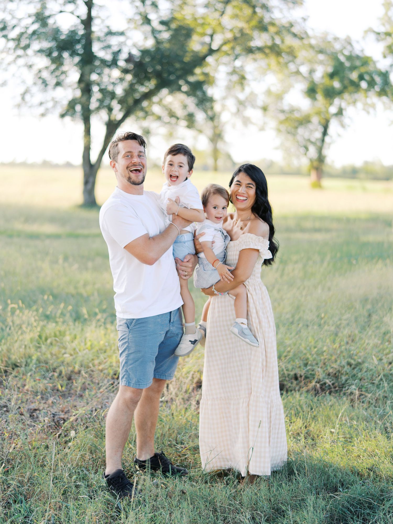Happy family of four posing in a sunny green field, parents holding two young children, wearing casual summer outfits.