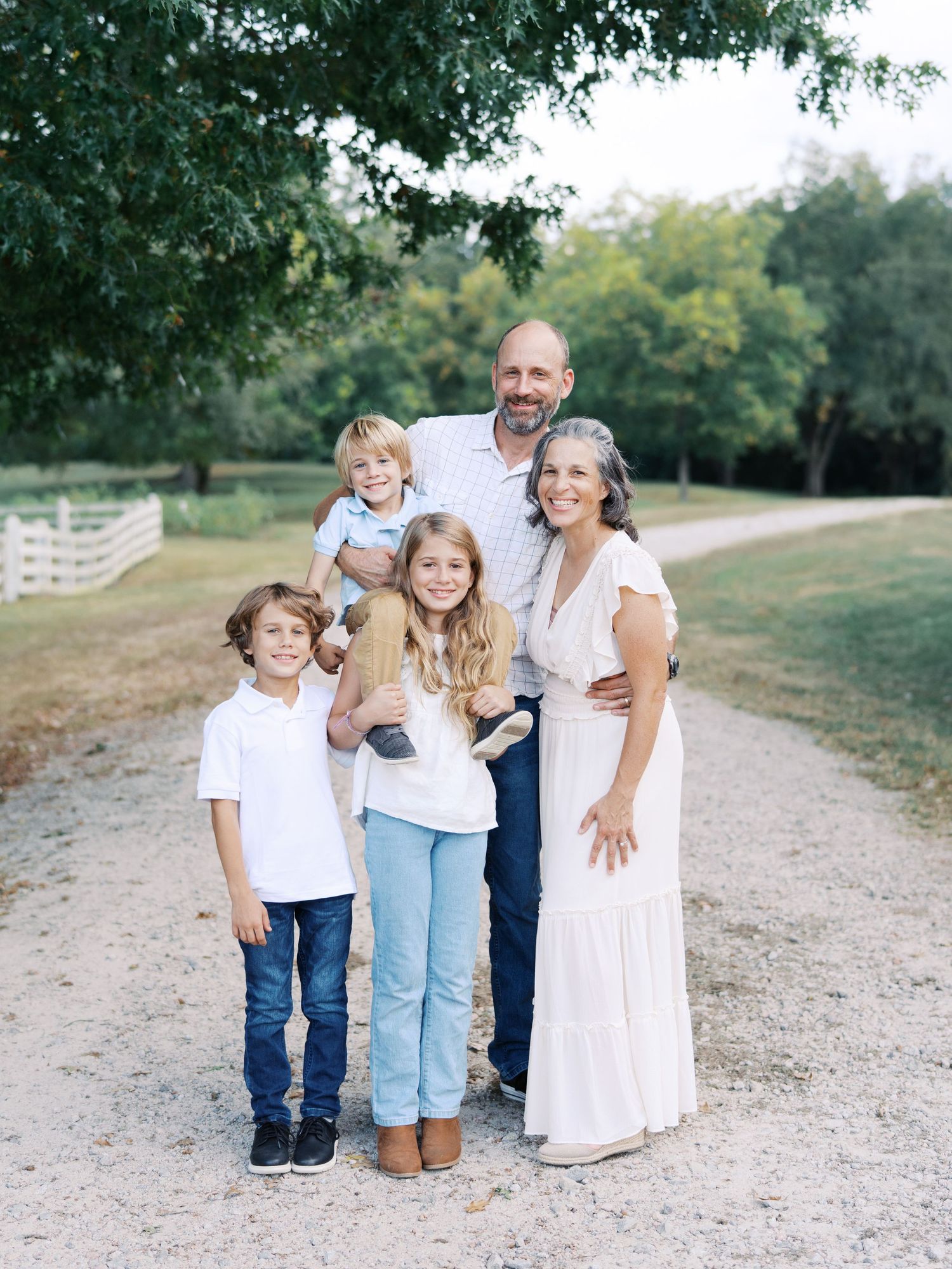 Family of five poses together on a gravel path surrounded by green trees in an outdoor setting.