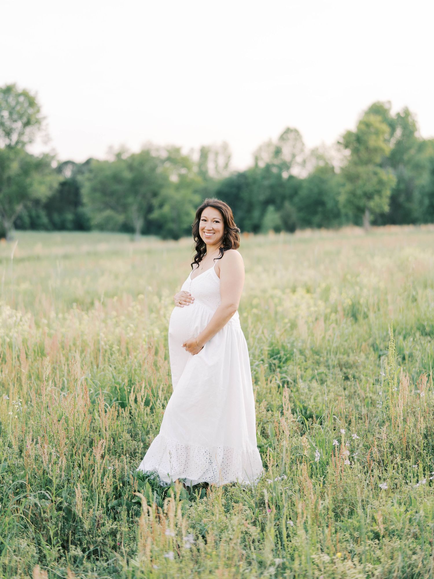 Expectant mother in white lace dress poses gracefully in a sunlit meadow with trees in the background.