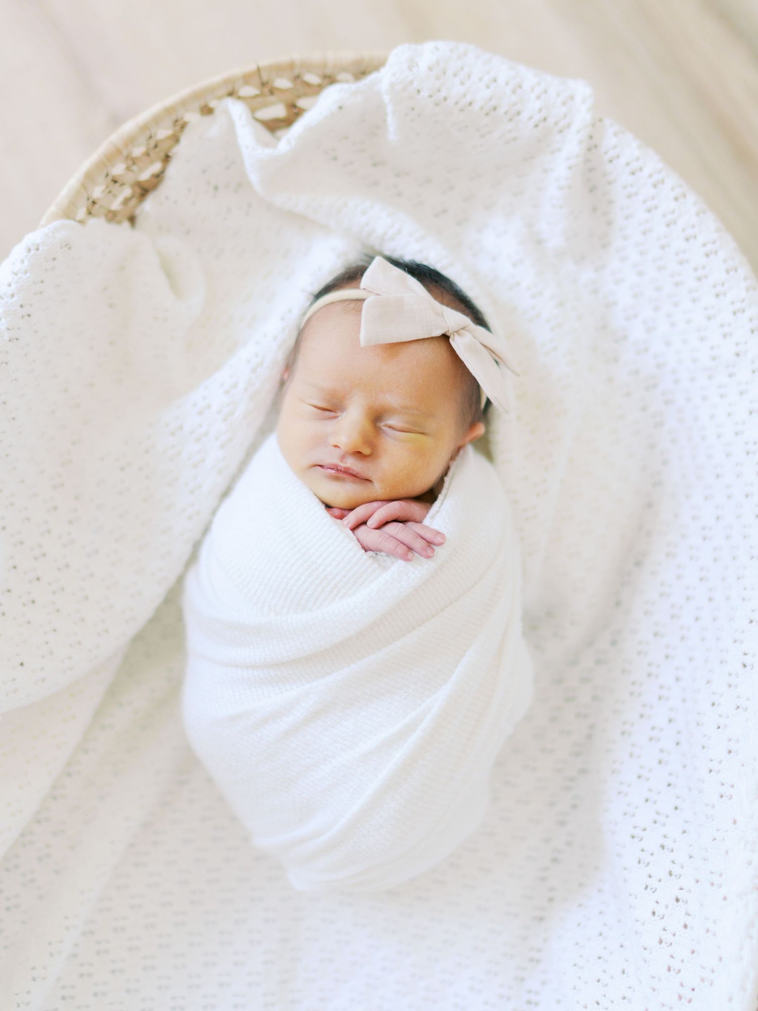 Sleeping newborn baby swaddled in white wrap with bow headband resting in wicker basket.