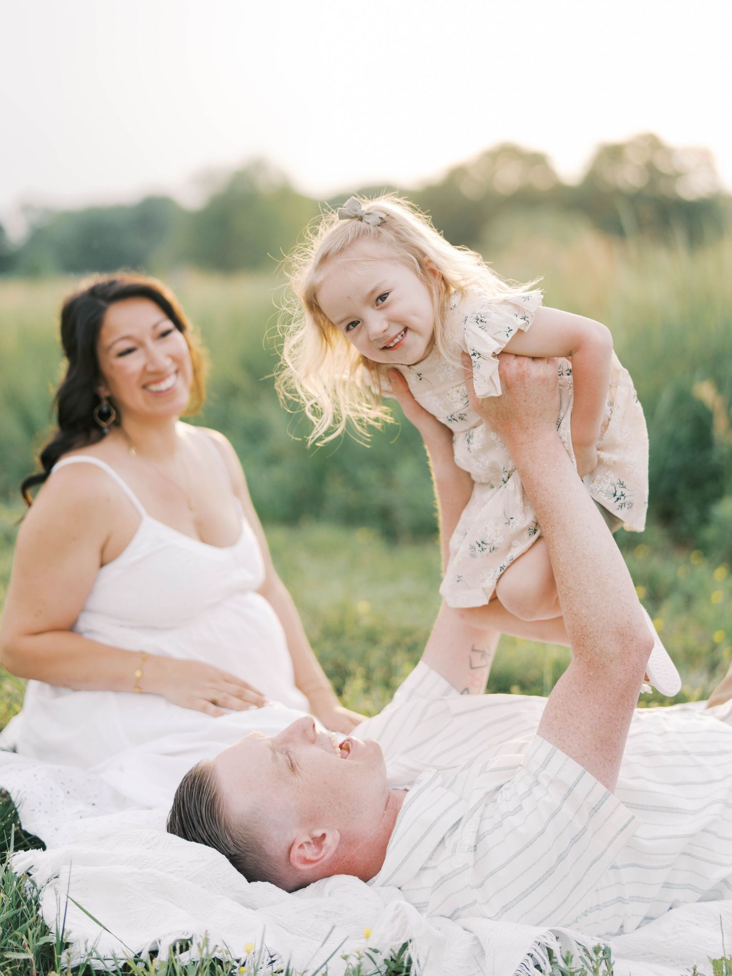 Playful family moment captured on blanket in sunny meadow during maternity photo session.