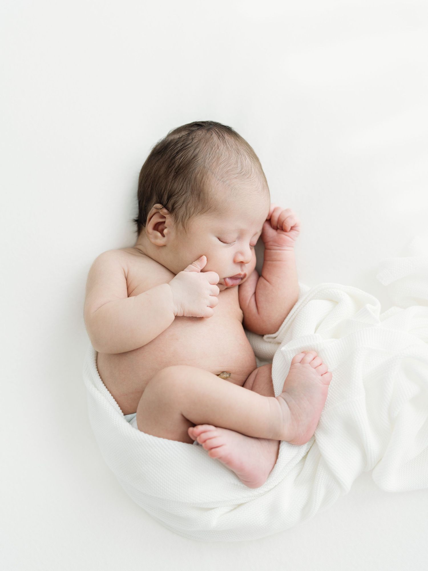 Sleeping newborn baby with dark hair curled up on a soft white blanket, hands near face, in a bright minimalist newborn photography style.
