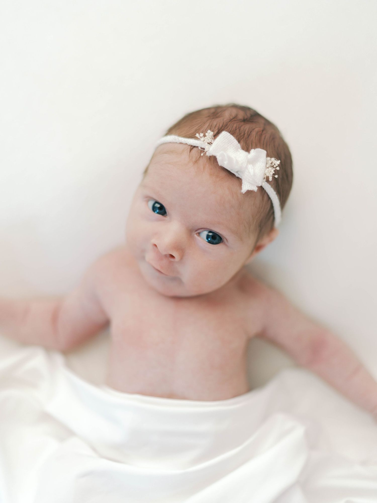 Newborn baby girl with blue eyes wearing a white bow headband, wrapped in a soft white blanket.
