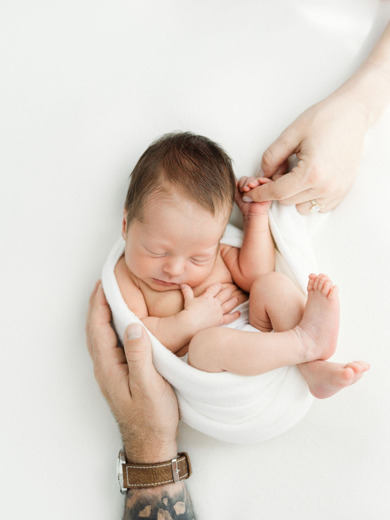 Newborn baby sleeping peacefully wrapped in white, cradled by two pairs of adult hands on a white background.