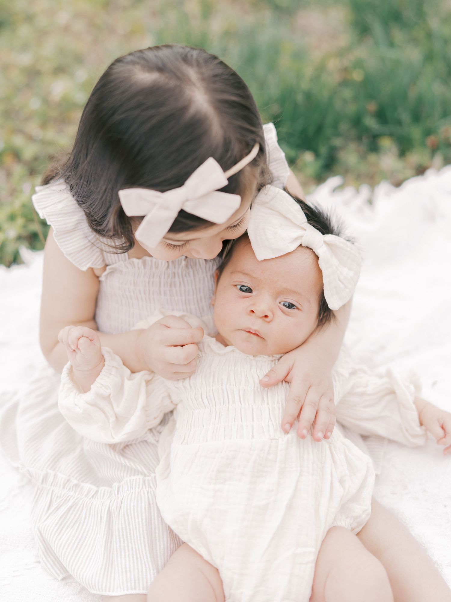 Two young sisters in white outfits and bow headbands sit together on a blanket outdoors, newborn cradled gently.