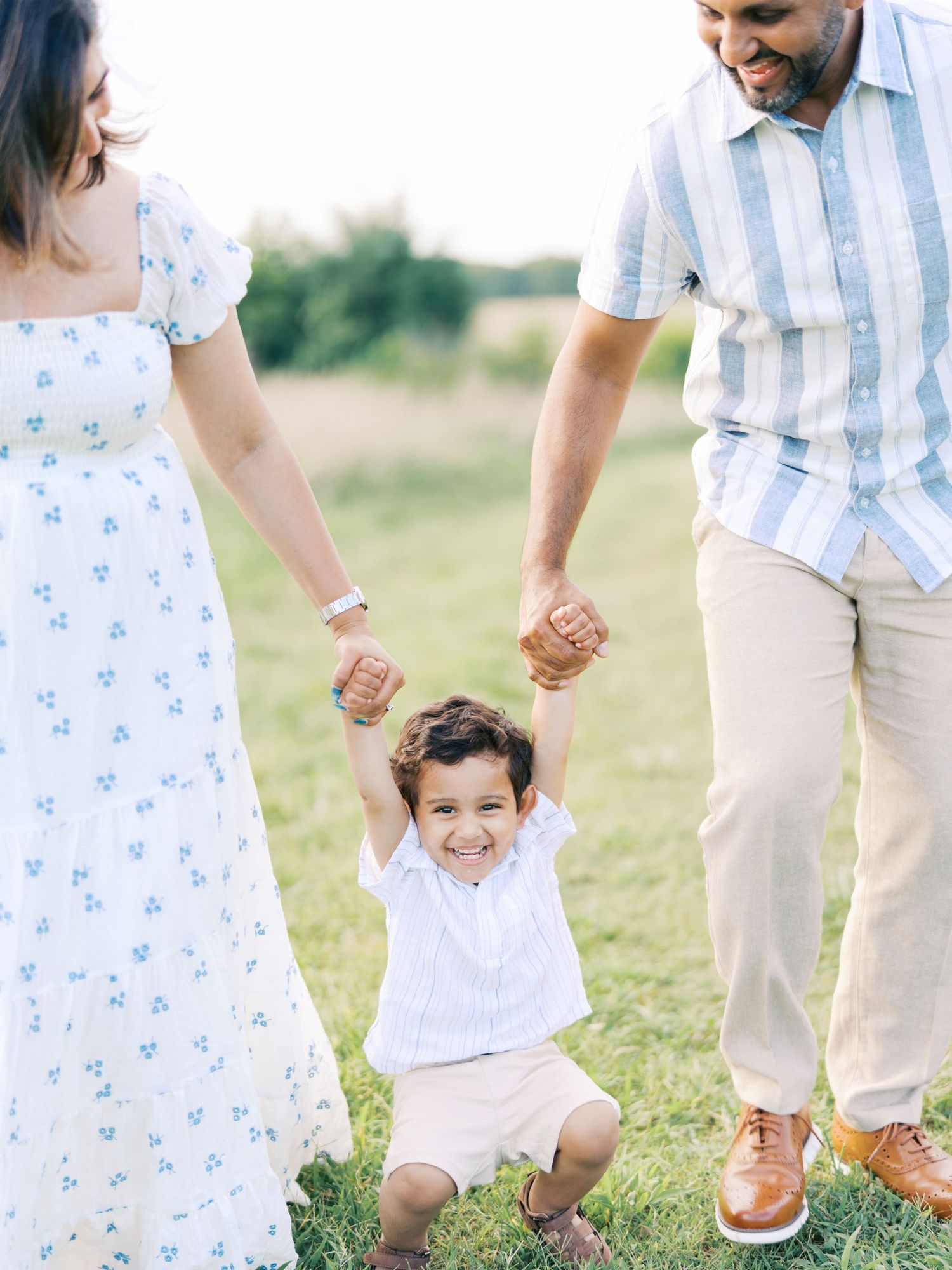 Family wearing coordinated white and blue outfits holds hands while walking together in a sunny outdoor field.
