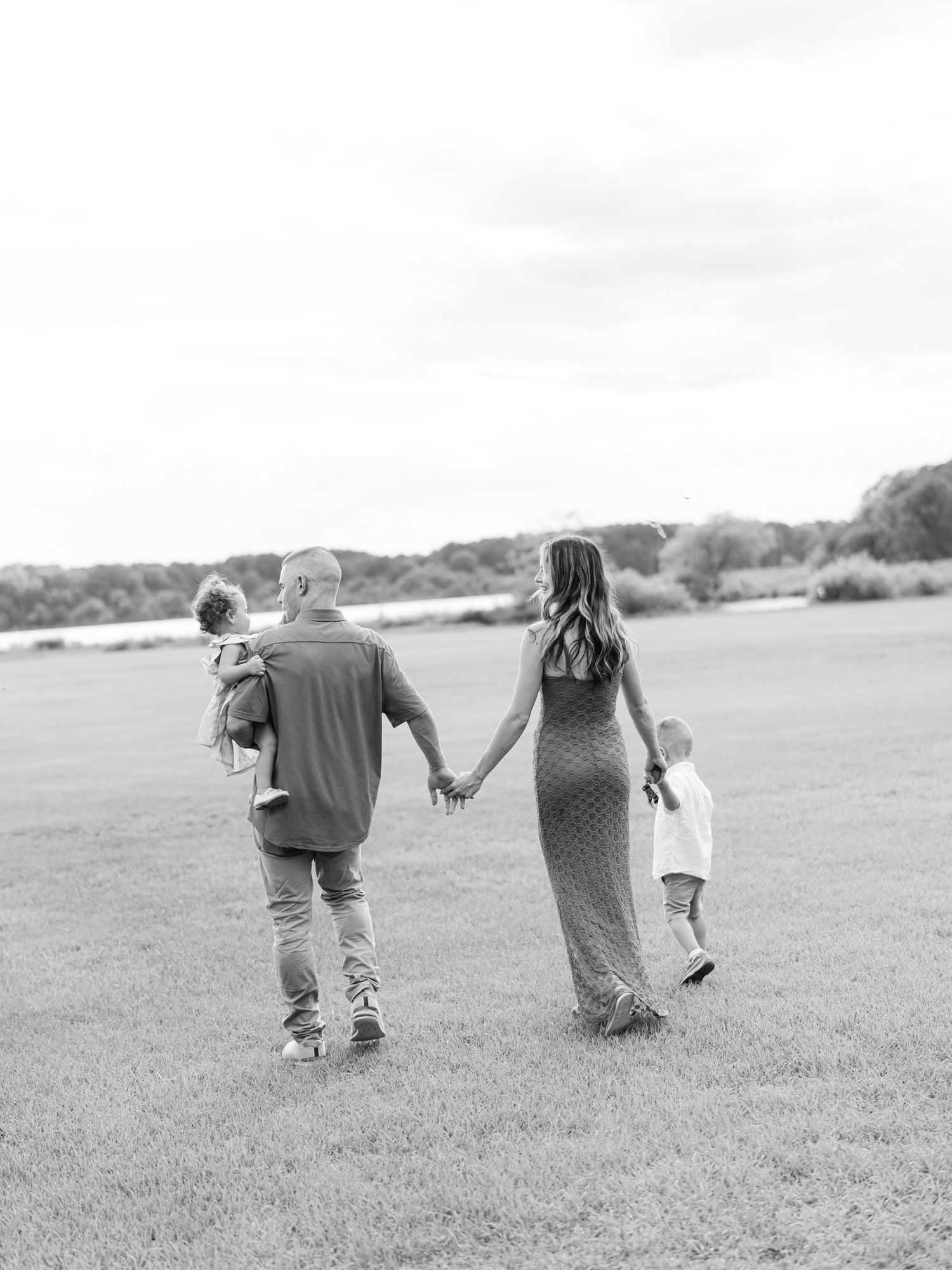 Black and white photo of a family holding hands walking through a grassy field toward a river.
