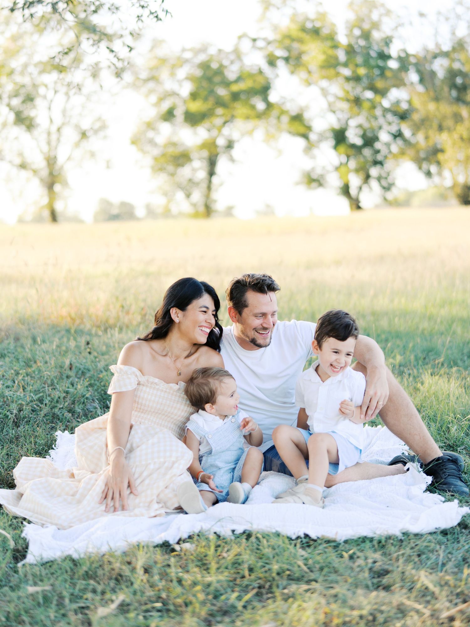 Family relaxes together on white blanket in grassy field during outdoor photoshoot.