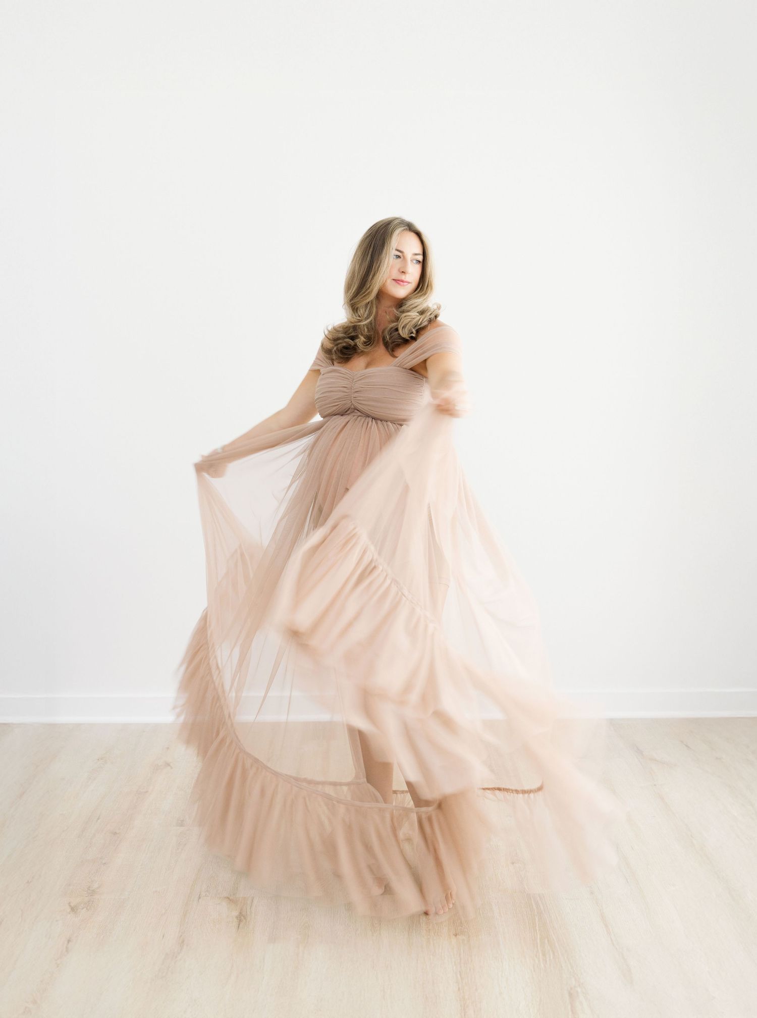 Woman in flowing blush tulle off-shoulder gown twirling gracefully in a bright white studio with light wood floors.