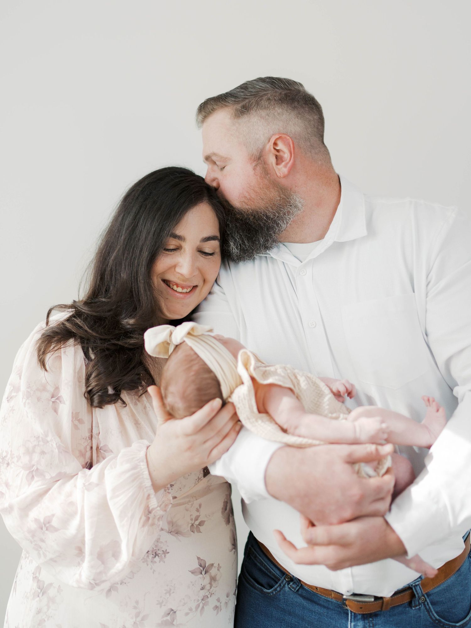 A couple smiles lovingly at their newborn baby, the father kissing the mother's head in a bright studio.