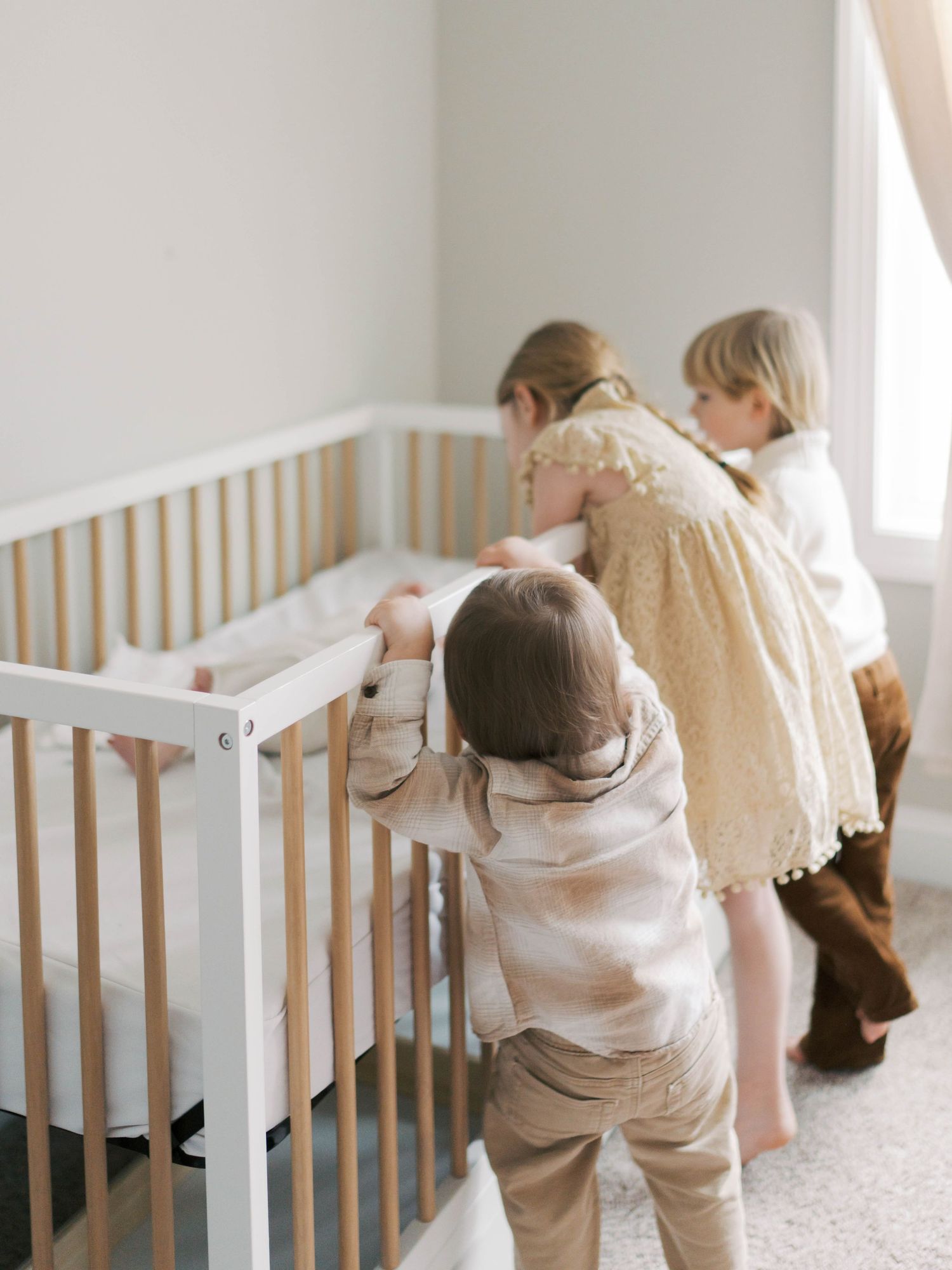 Three young children leaning over a white and wood crib, curiously looking at a newborn baby inside.