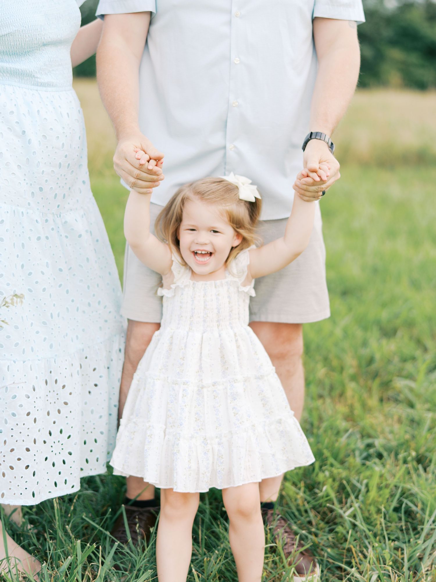 A child in a white dress holds hands with adults while playing in a grassy field on a sunny day.