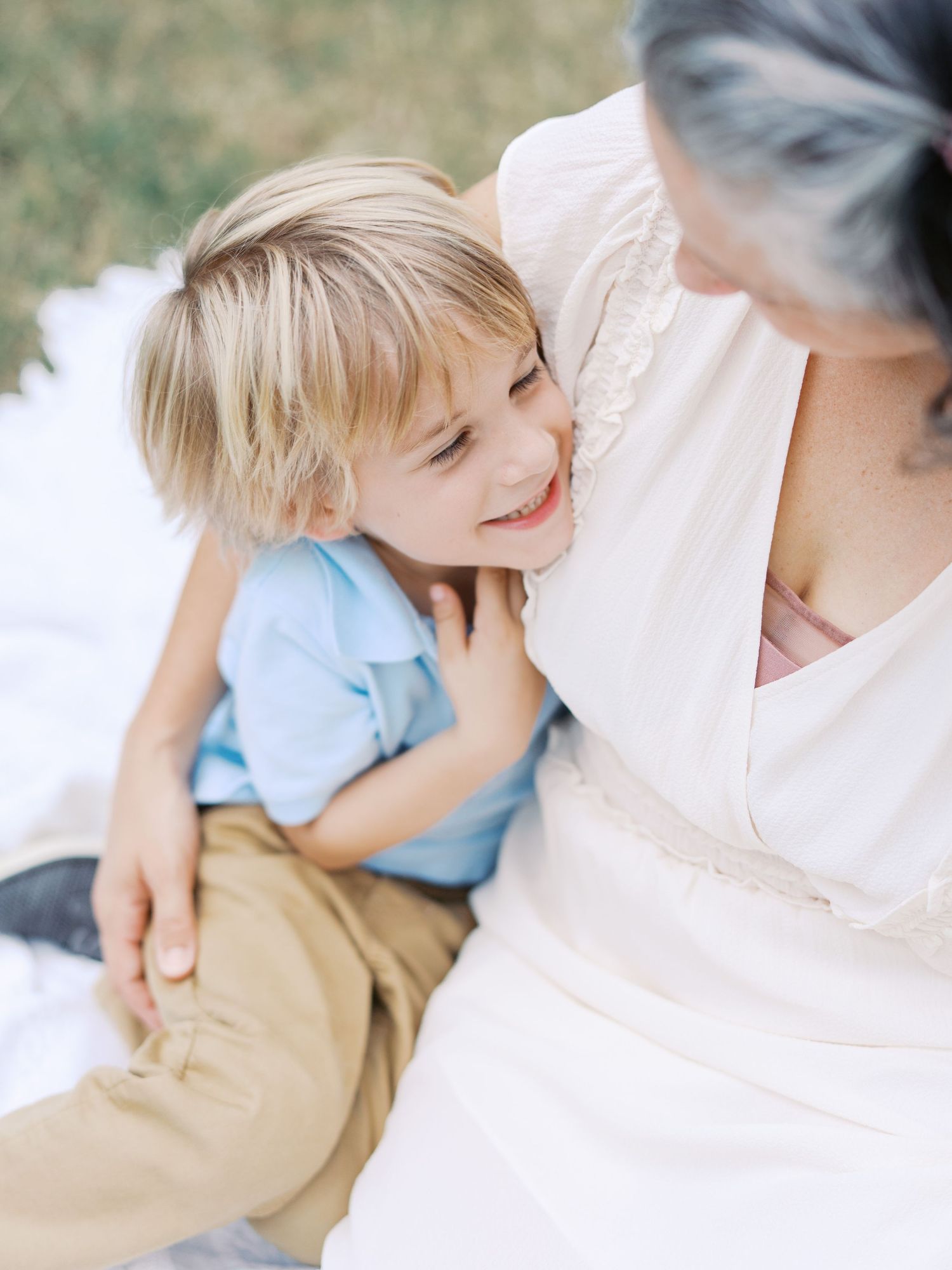 A young child in a blue shirt smiles joyfully while sitting close to someone wearing white in an outdoor setting.