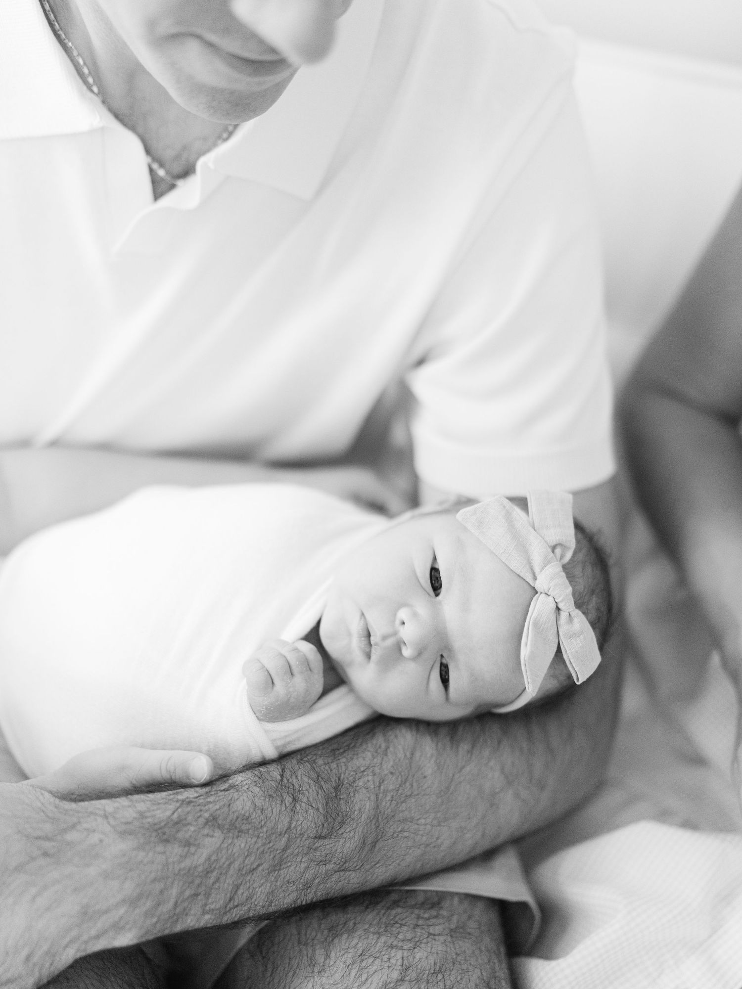 Black and white photo of a newborn baby girl with bow headband cradled in her fathers arms.