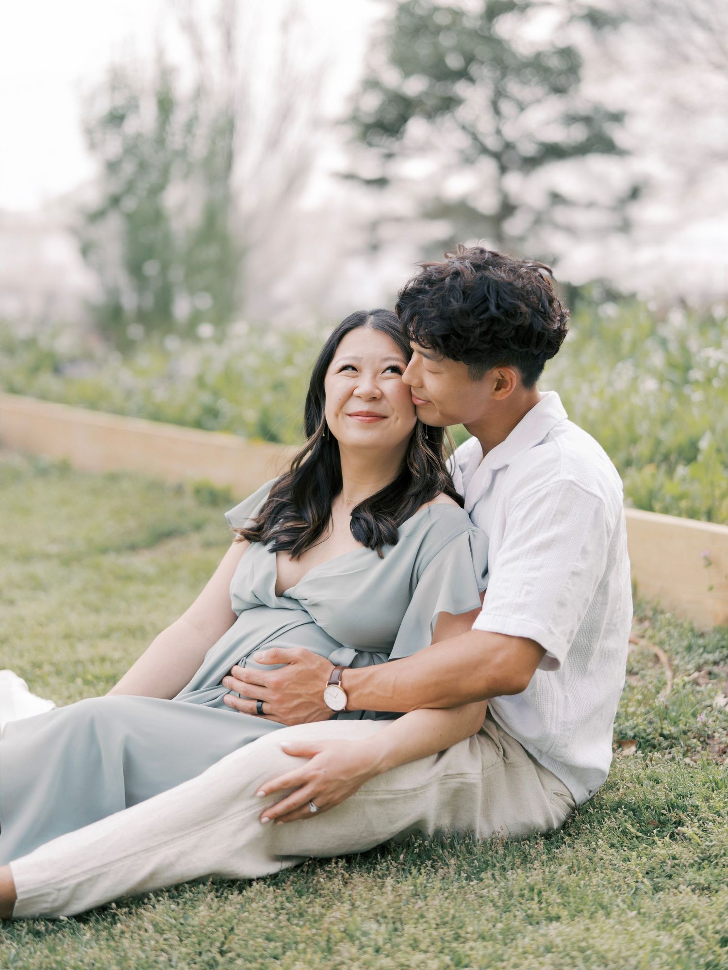 A couple relaxes on grass during their outdoor maternity photoshoot with spring blossoms in background.