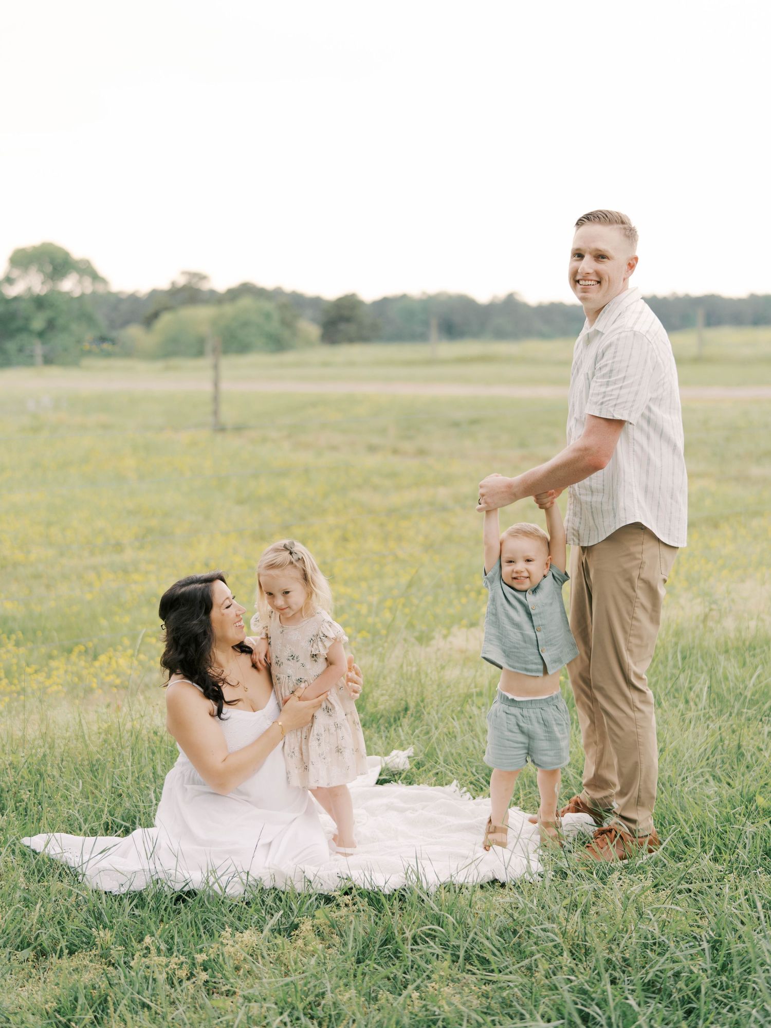 Family enjoying a playful moment together on a white blanket in a sunny meadow with wildflowers.