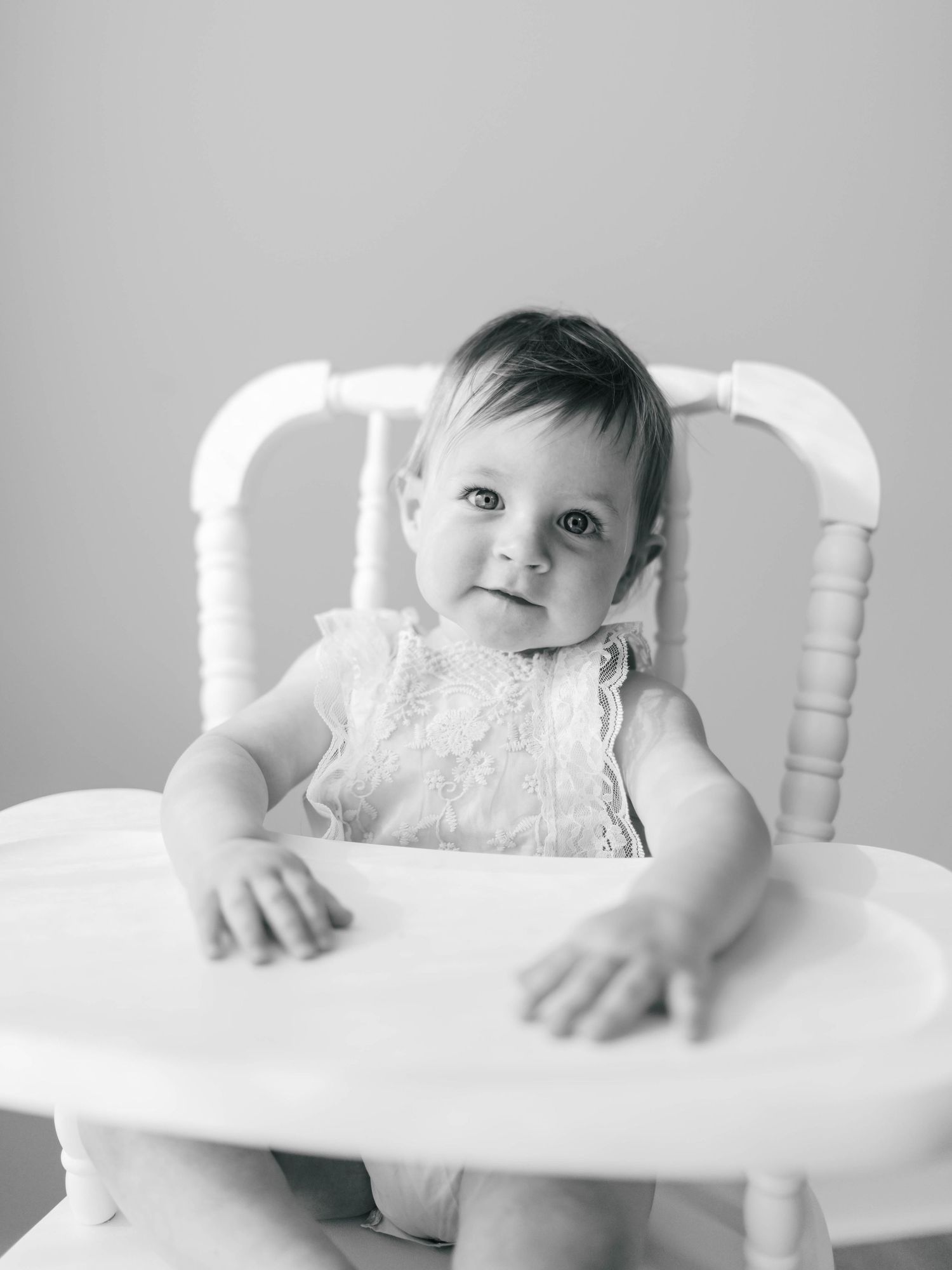 Black and white photo of a cute baby girl sitting in a white high chair, smiling at the camera.