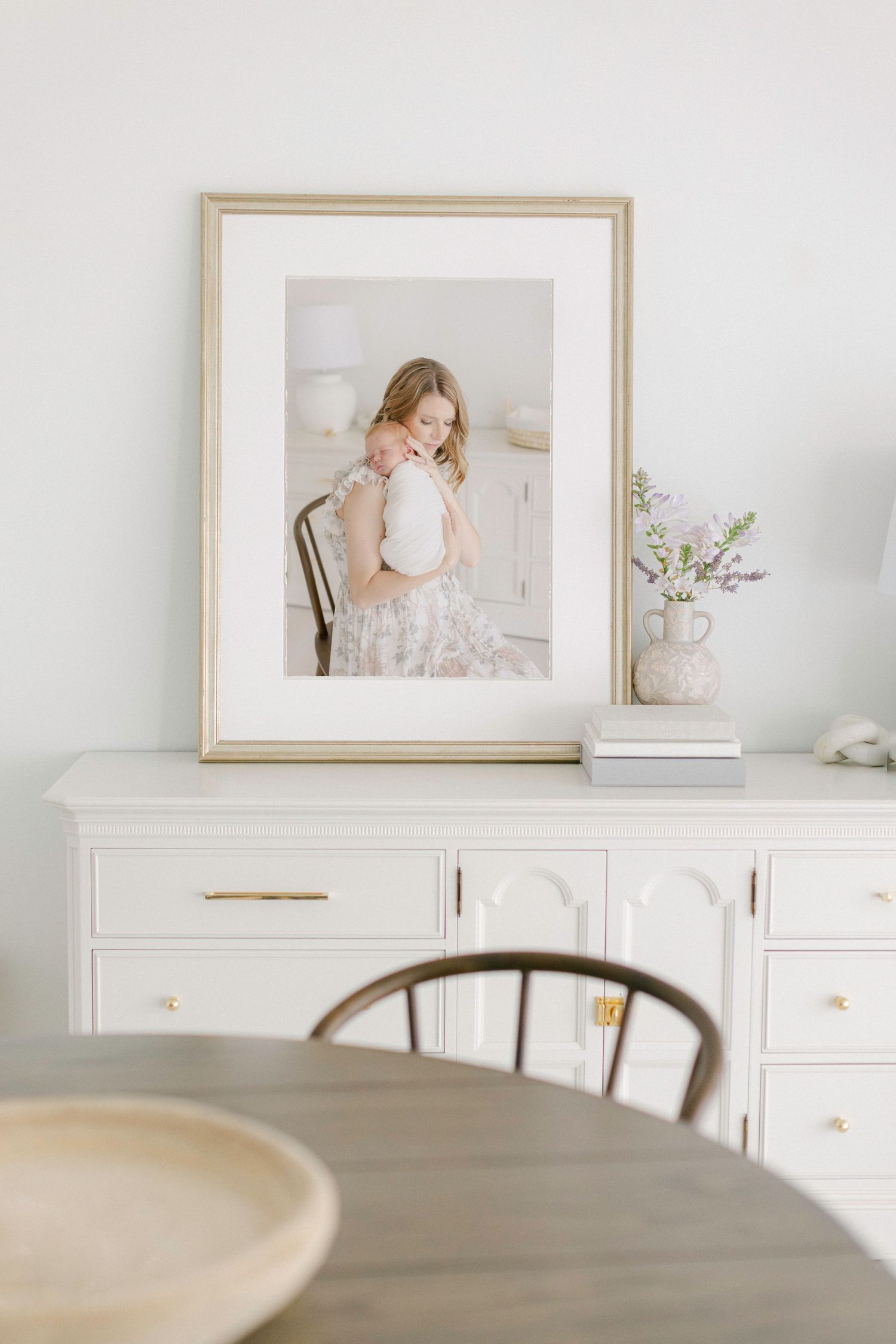 Framed portrait of mother holding baby displayed on white sideboard with gold hardware, beside a floral vase in a bright dining room.
